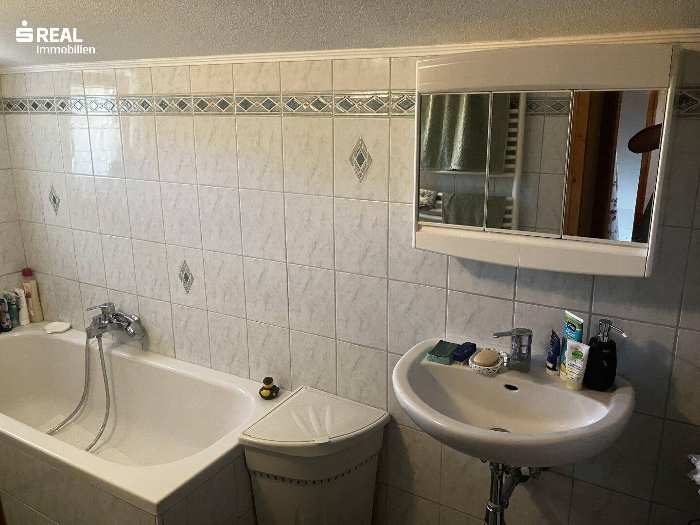 Badezimmer mit Badewanne, Waschbecken und hellen Fliesen im 80er-Jahre-Stil.