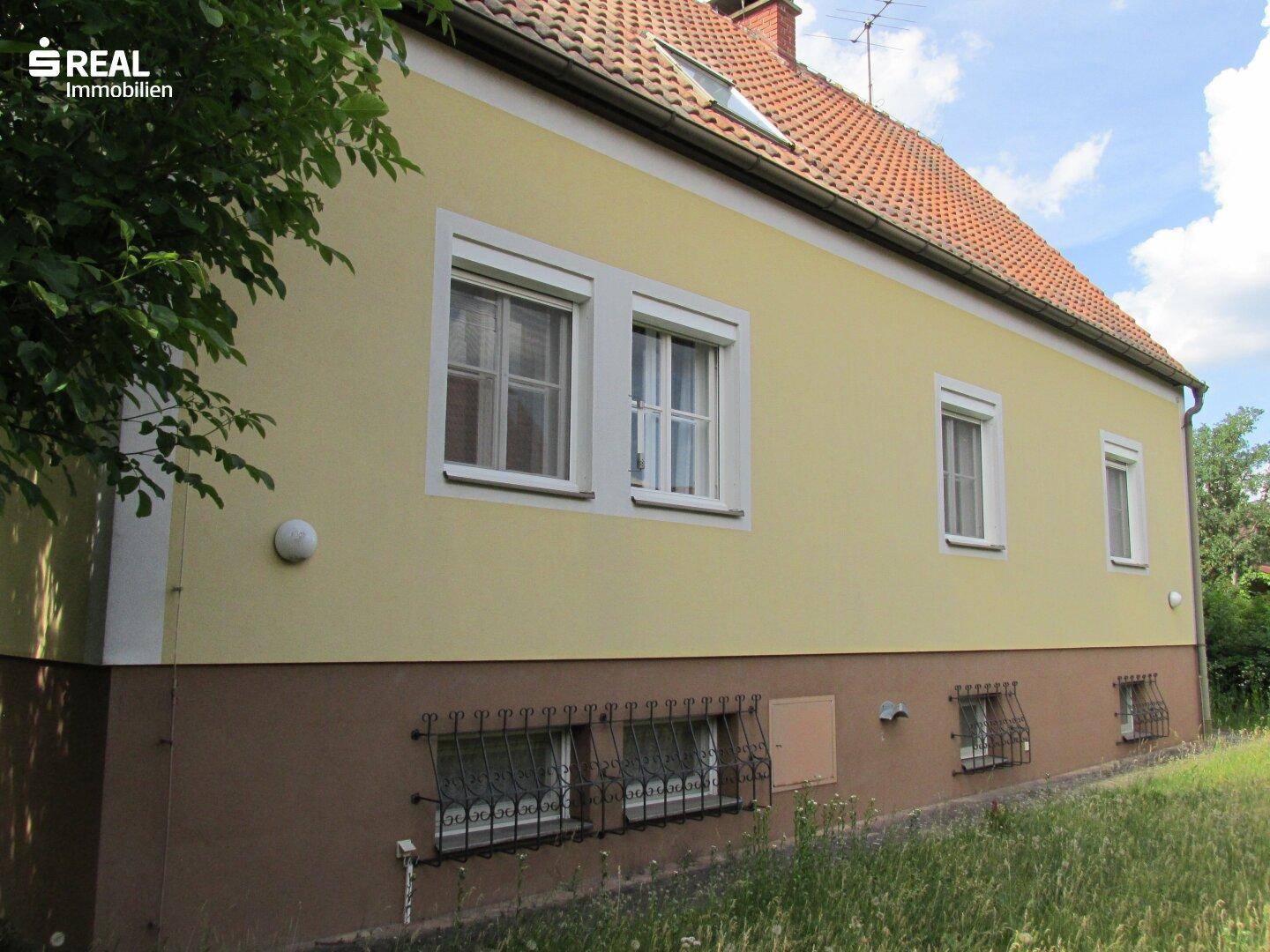 Seitenansicht des Hauses mit gelber Fassade, Rollläden und vergitterten Kellerfenstern.