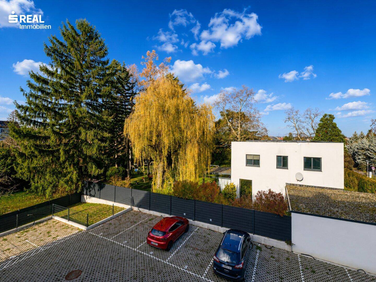 Außenparkplätze mit Schotterbelag, umgeben von Bäumen und einem Zaun, mit Blick auf ein Gebäude.