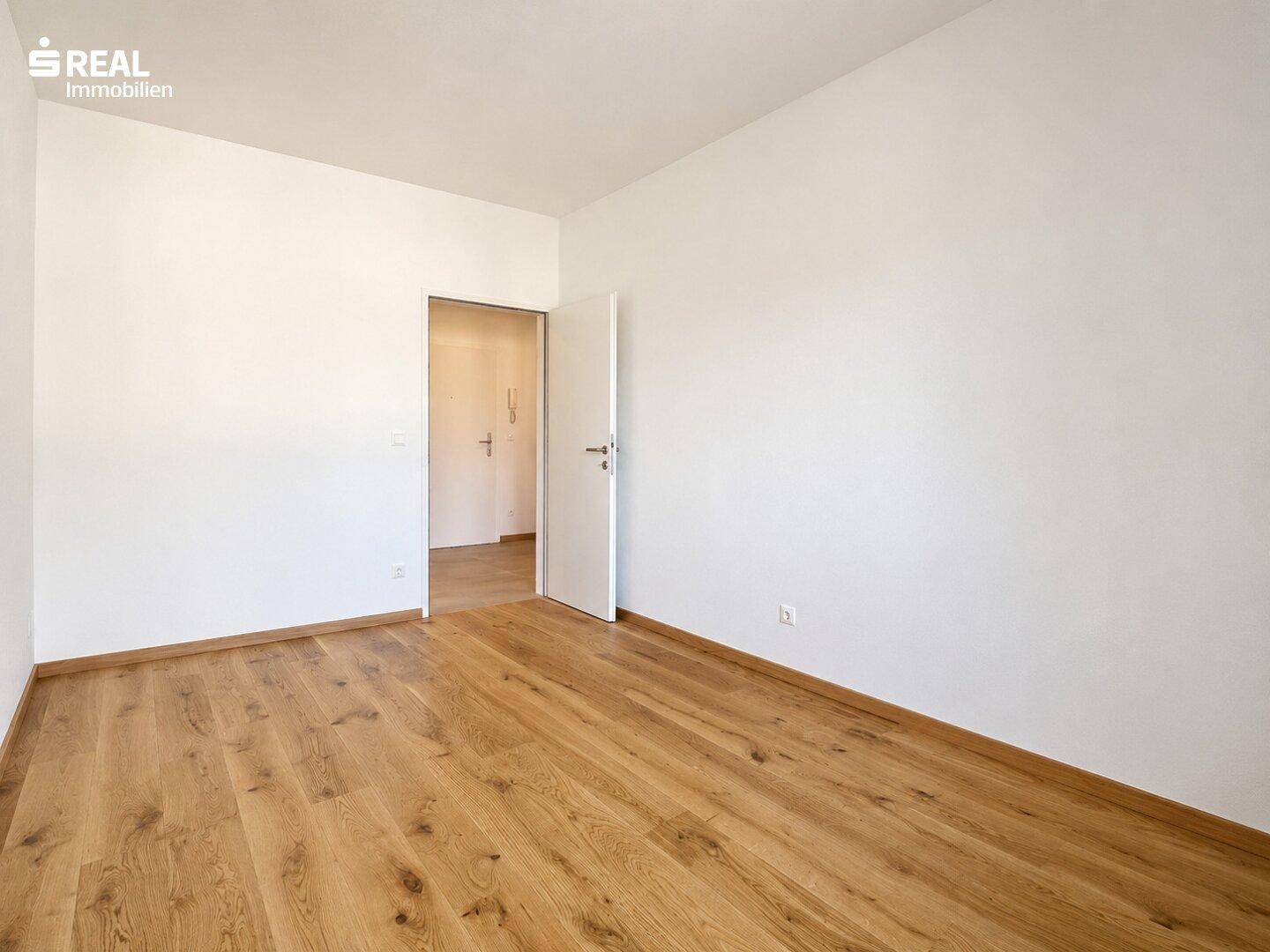 Leeres Zimmer mit warmem Holzboden und weißen Wänden, ideal für Schlaf- oder Arbeitszimmer.