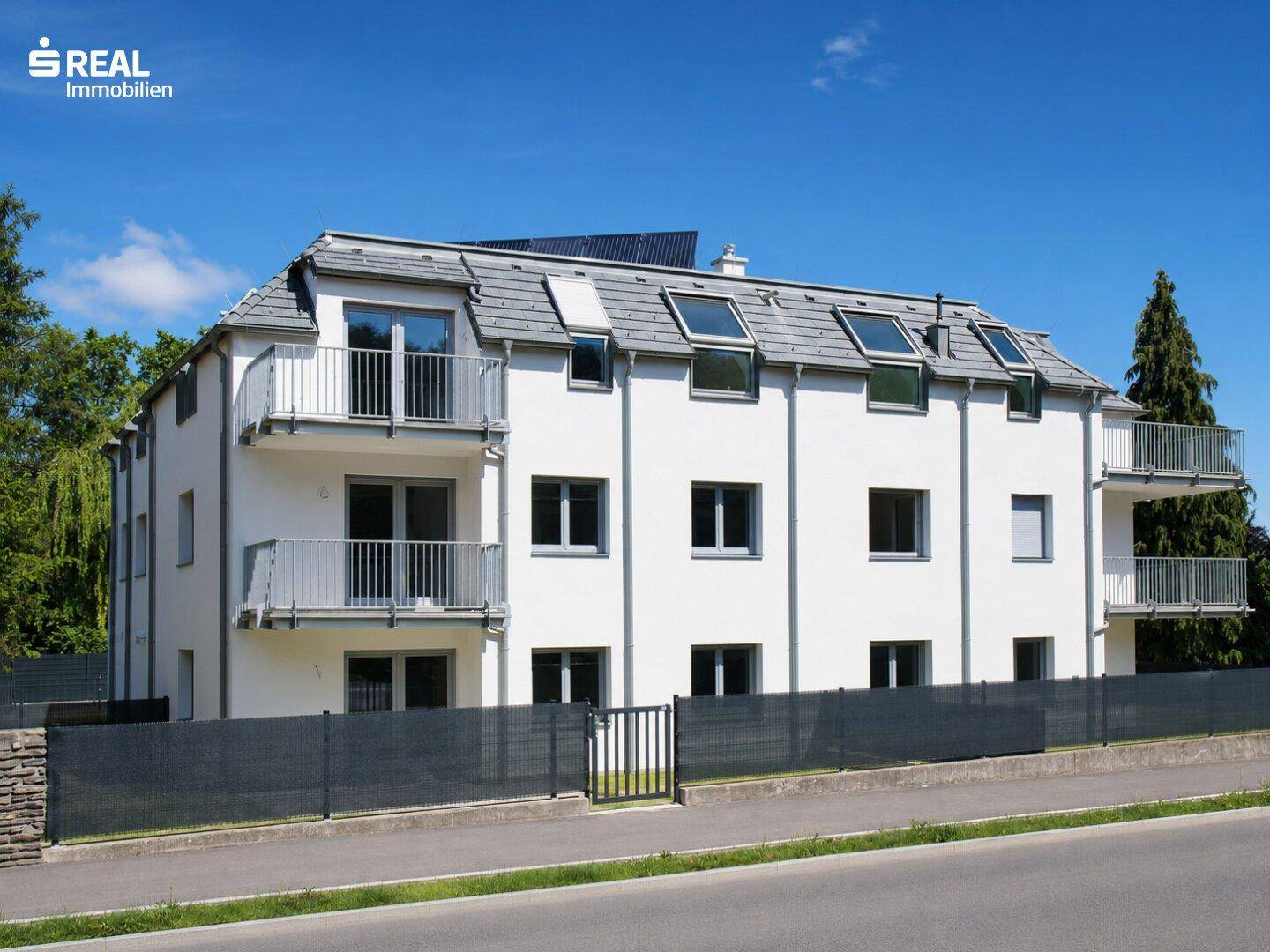 Modernes Mehrfamilienhaus mit weißer Fassade, Balkonen und Dachfenstern unter blauem Himmel.