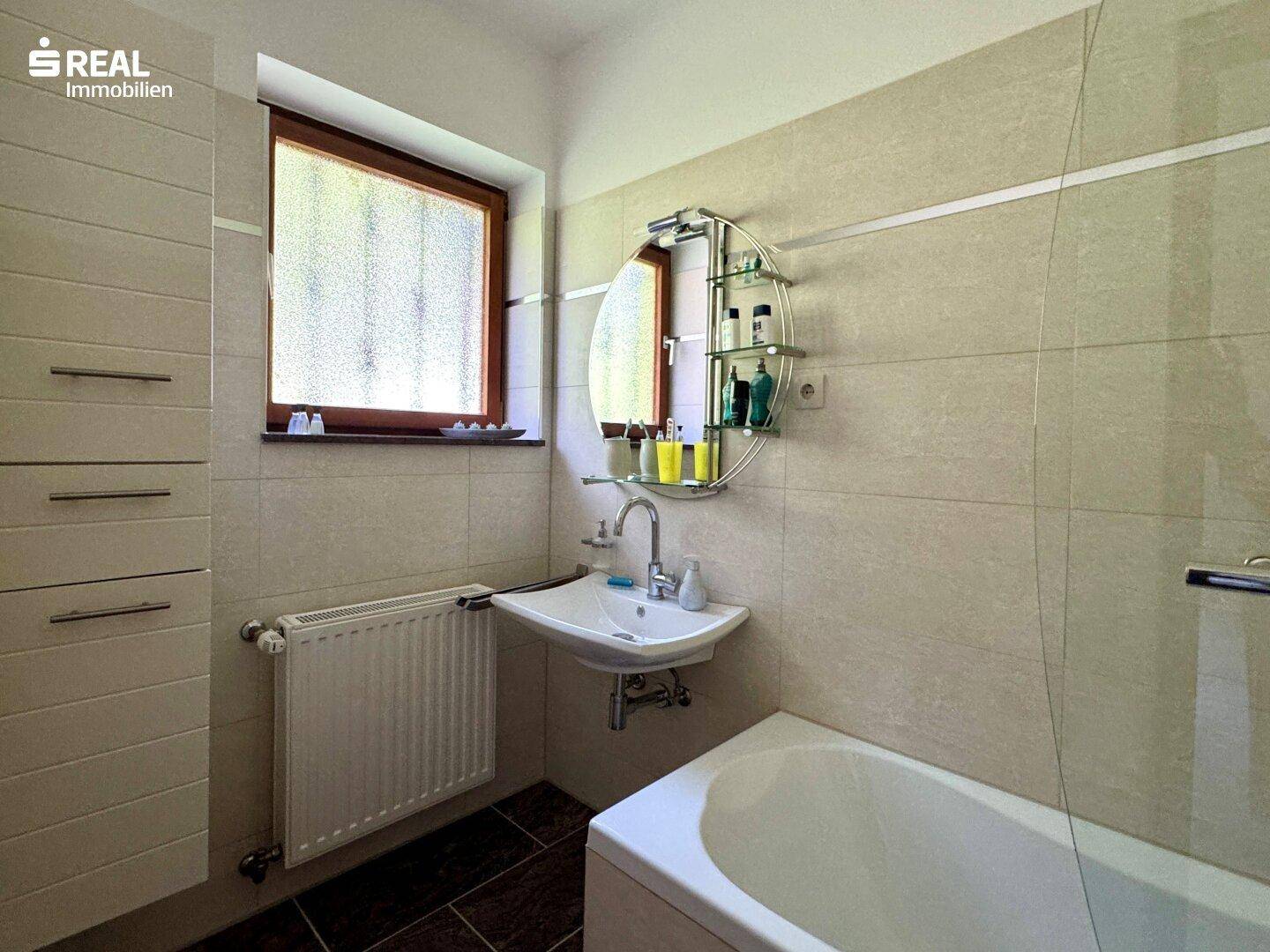 Helles Badezimmer mit Badewanne, Waschbecken und Fenster, ausgestattet mit beigen Fliesen.