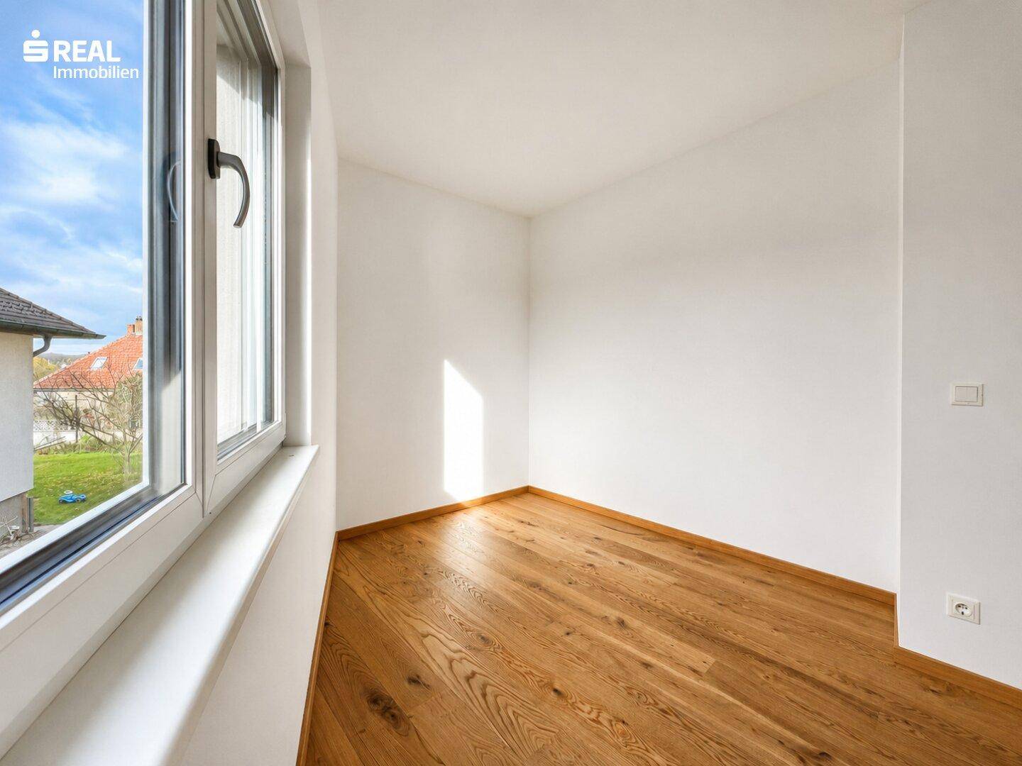 Lichtdurchflutetes Zimmer mit hellem Holzboden und großem Fenster, ideal als Schlafzimmer.