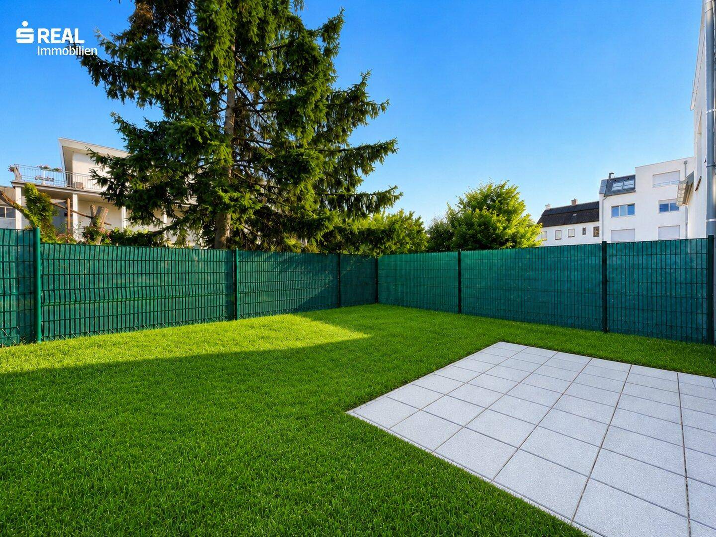 Gepflegter Gartenbereich mit Rasenfläche und einer kleinen gepflasterten Terrasse, umgeben von einem grünen Zaun.