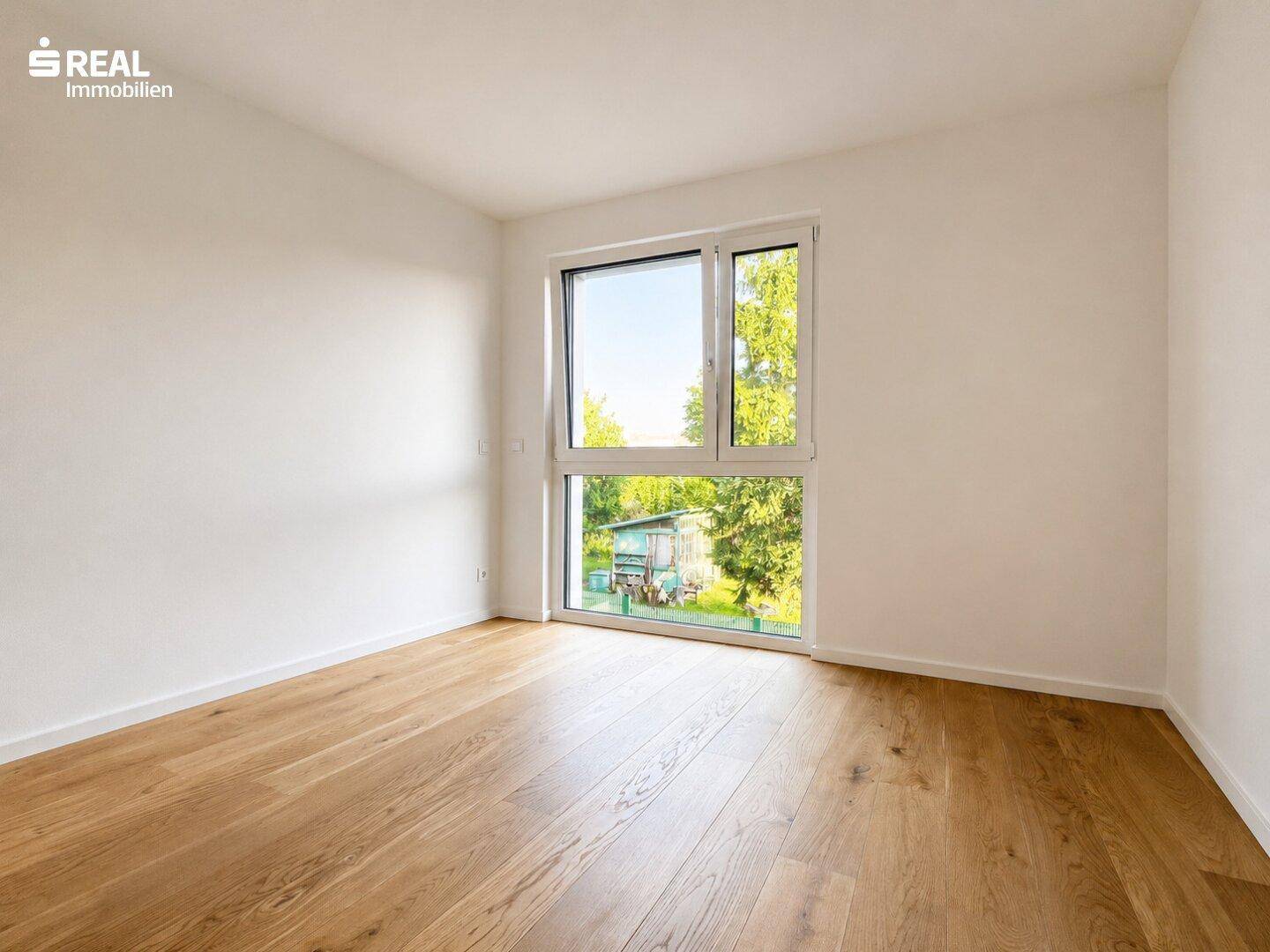 Geräumiges Schlafzimmer mit Holzboden und großem Fenster, das viel natürliches Licht und eine grüne Aussicht bietet.