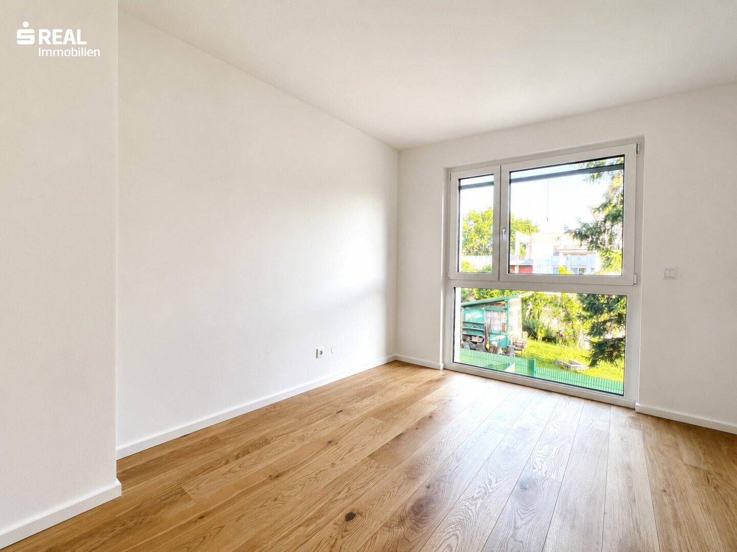 Lichtdurchflutetes Schlafzimmer mit Holzboden und großem Fenster, das einen angenehmen Blick ins Grüne ermöglicht.