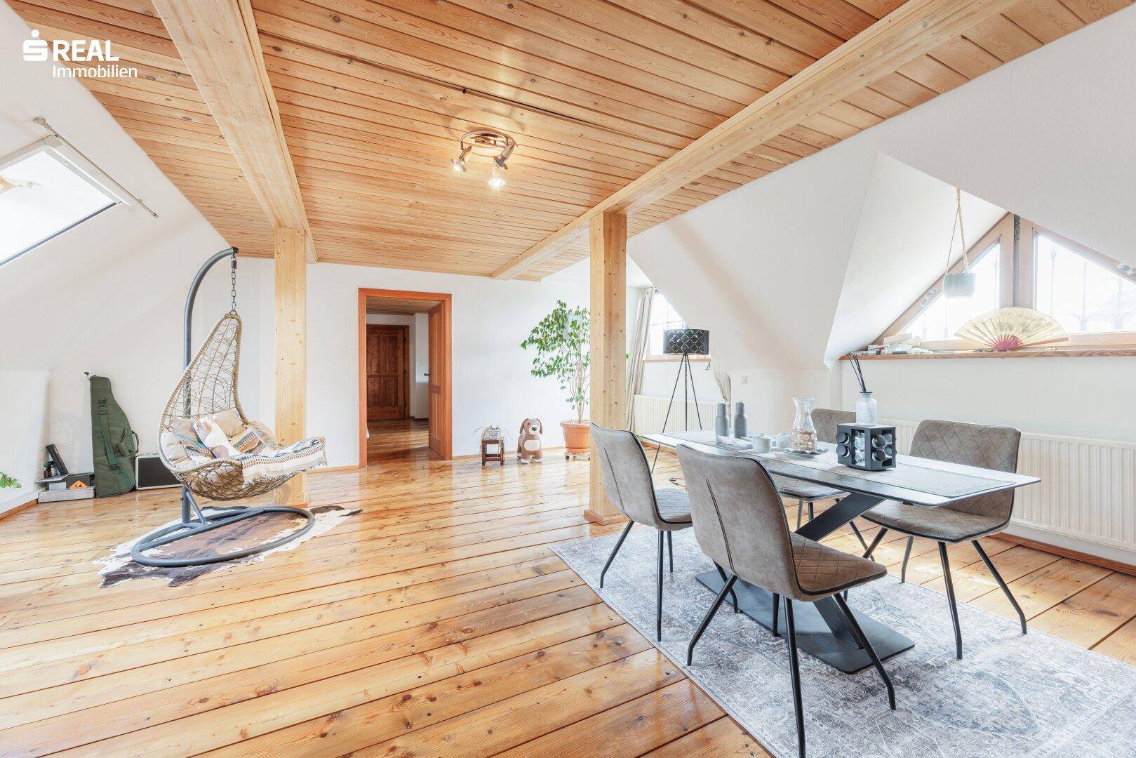 Essbereich im offenen Dachgeschoss mit Holzboden, Holzbalken und viel natürlichem Licht durch große Fenster.