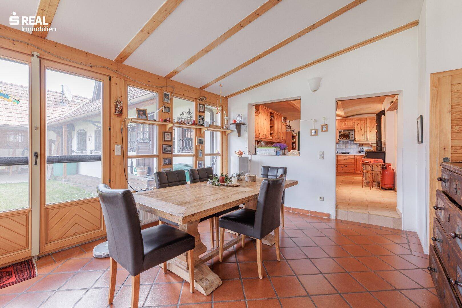 Essbereich im Wintergarten mit Holztisch und Zugang zu einer traditionellen Küche mit Holzelementen.