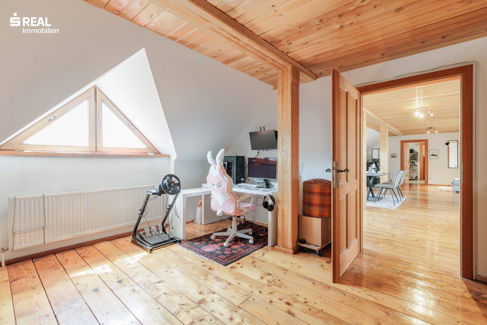 Gemütliches Arbeitszimmer im Dachgeschoss mit Holzboden, Holzbalken und dreieckigem Fenster.