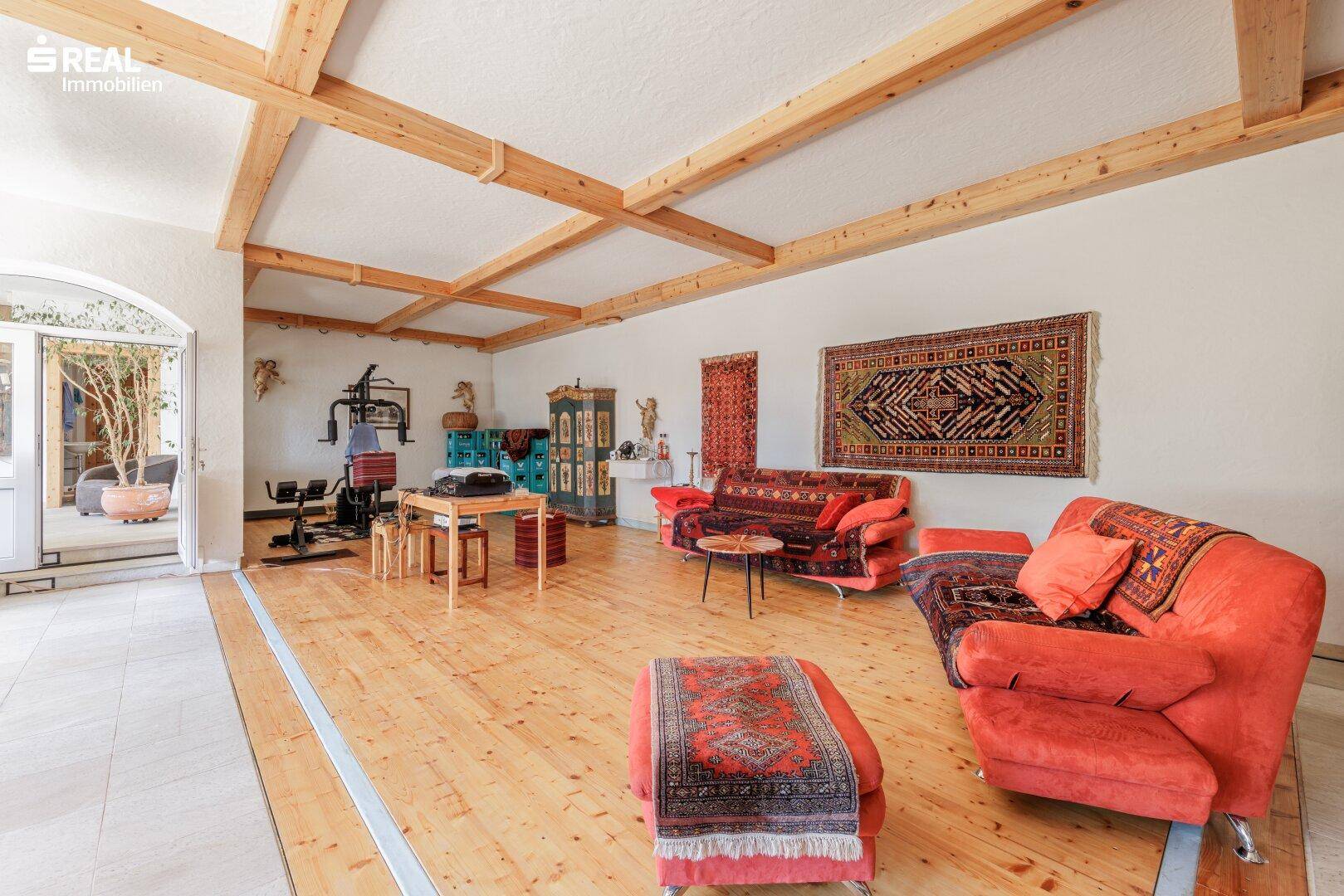 Großes Wohnzimmer mit Holzdielenboden, roten Sofas, Fitnessgeräten und Zugang zu einem Außenbereich.