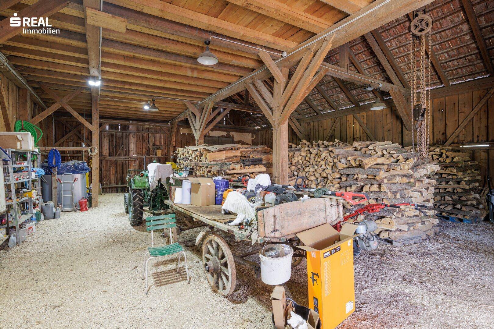 Geräumige Scheune mit Holzbalken, landwirtschaftlichen Geräten und großem Holzstapel.