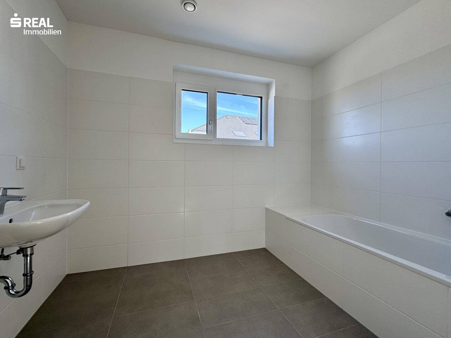 Modernes Badezimmer mit weißen Fliesen, Badewanne und Waschbecken unter einem Fenster.