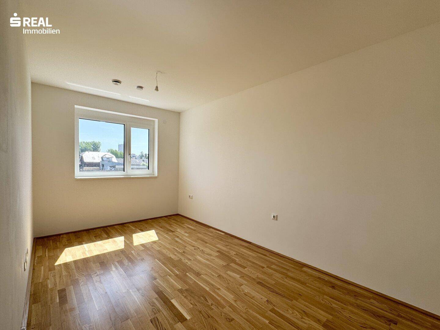 Helles Schlafzimmer mit Parkettboden und Fenster, das viel Tageslicht hereinlässt.
