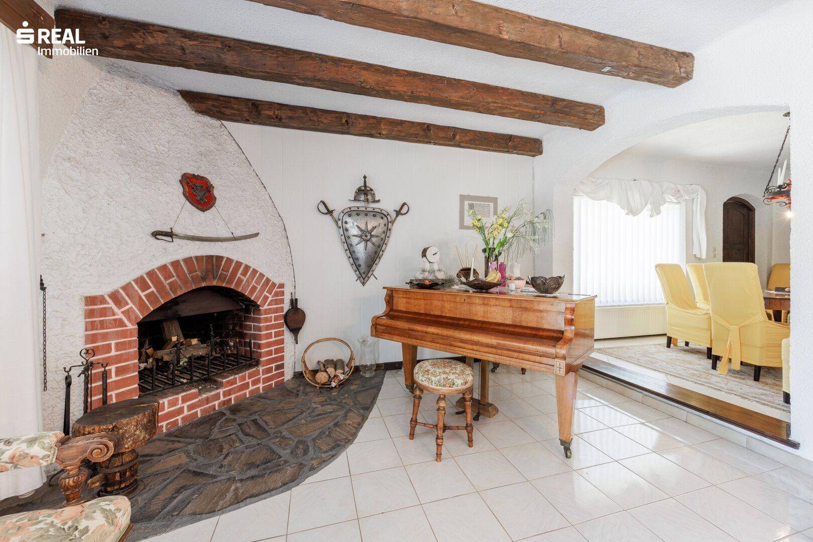 Wohnzimmer mit großem gemauertem Kamin, freiliegenden Holzbalken und einem Klavier.