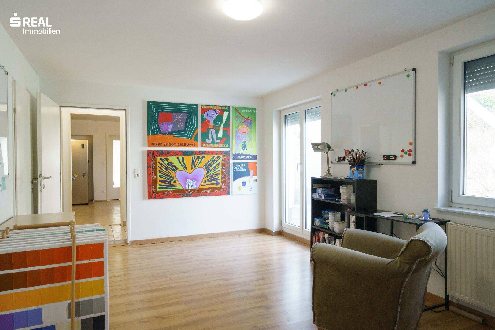 Geräumiger Wohnbereich mit großen Fenstern, Holzboden und farbenfrohen Kunstwerken an den Wänden.