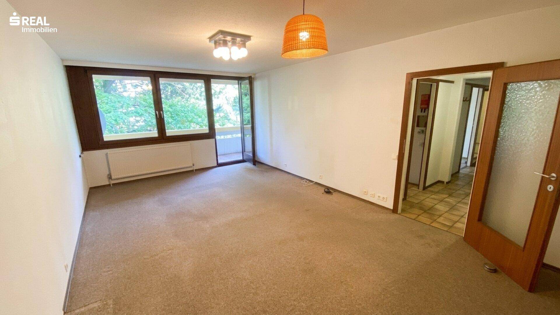 Leeres Wohnzimmer mit Teppichboden, großem Fenster und Zugang zum Balkon.