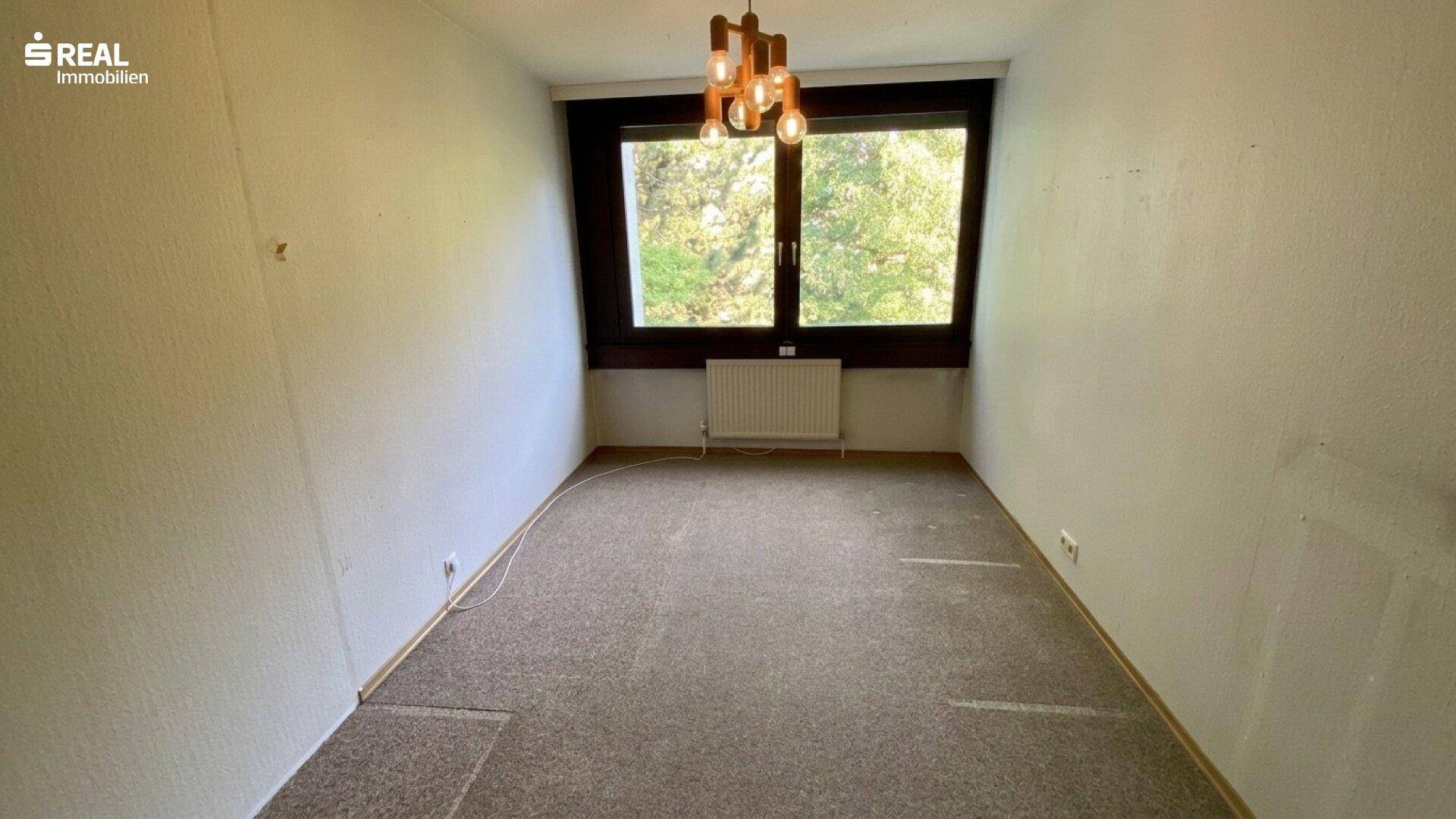 Leeres Zimmer mit Teppichboden und großem Fenster, das viel Tageslicht hereinlässt.