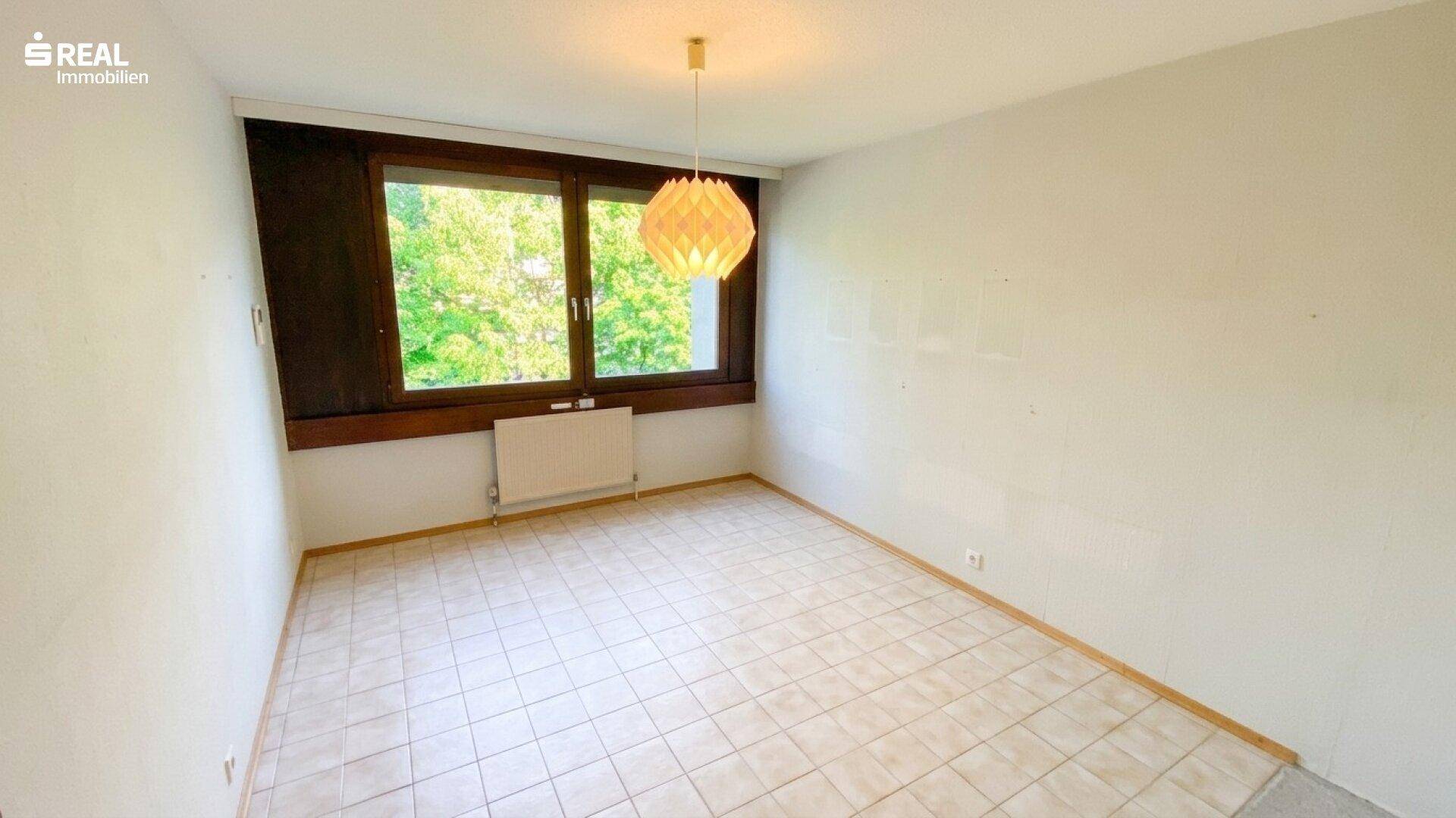 Leeres Zimmer mit hellen Fliesen, großem Fenster und einer auffälligen Deckenleuchte.