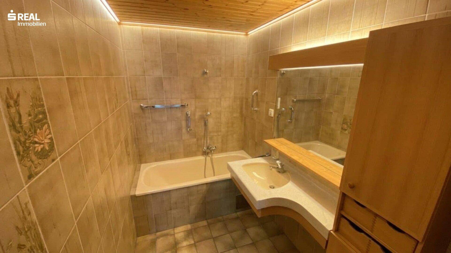Gefliestes Badezimmer mit Badewanne, Waschbecken und Holzdecke mit indirekter Beleuchtung.