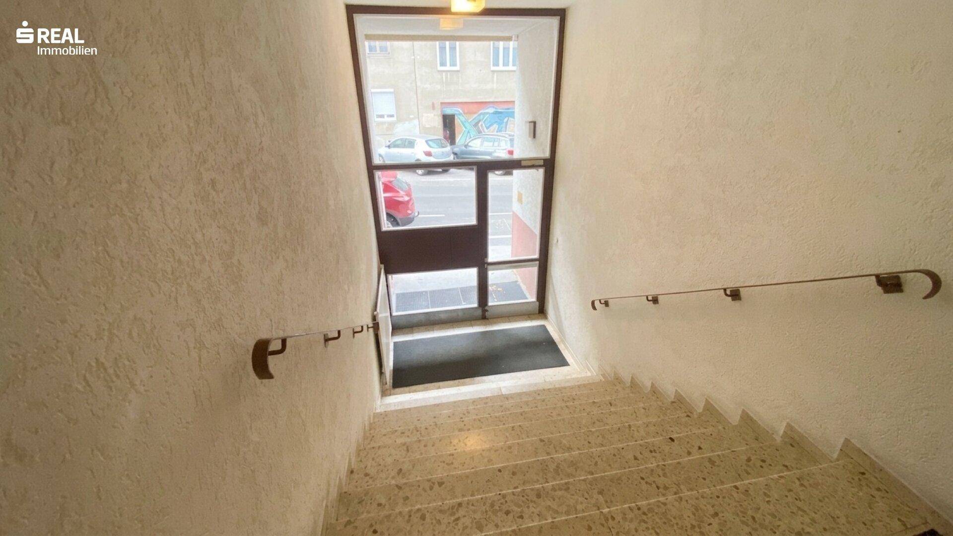 Treppenhaus mit Handläufen und Ausgangstür, die zur Straße führt, in einem Mehrfamilienhaus.
