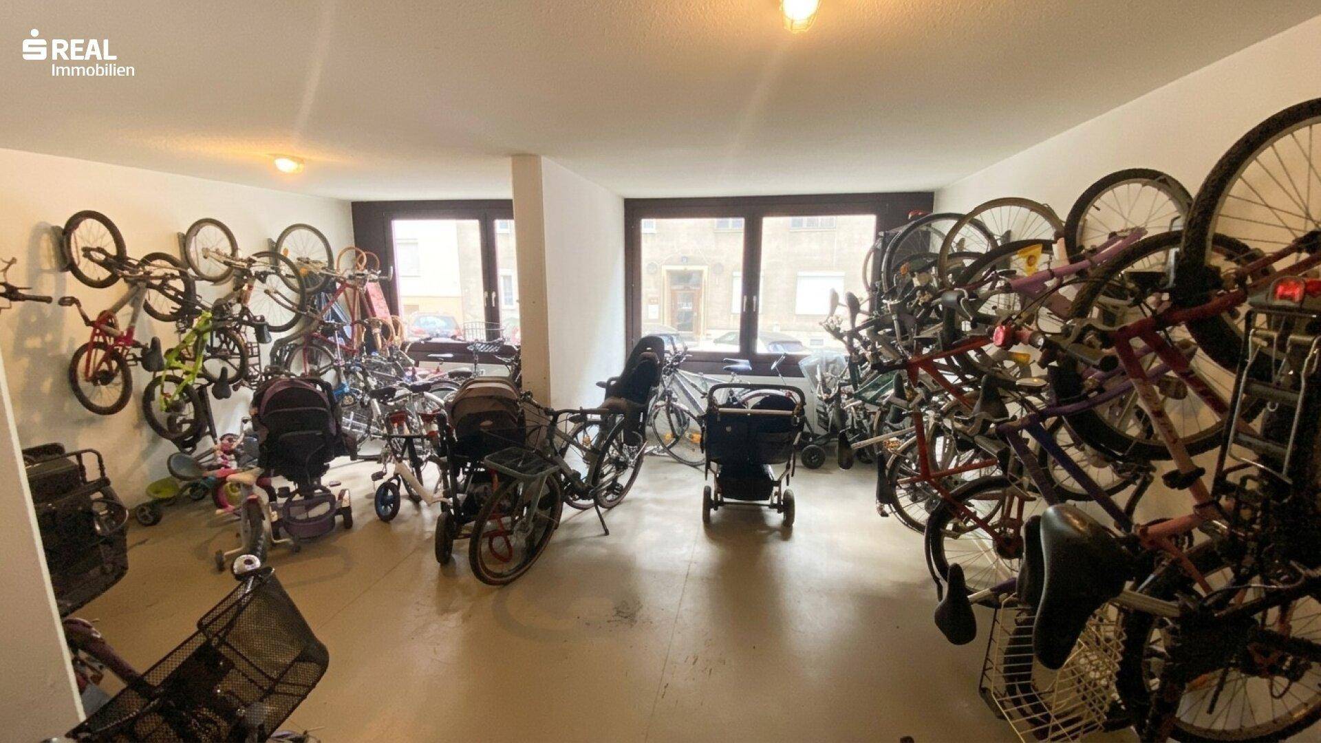 Gemeinschaftlicher Fahrradraum mit vielen Fahrrädern und Kinderwagen, gut beleuchtet.