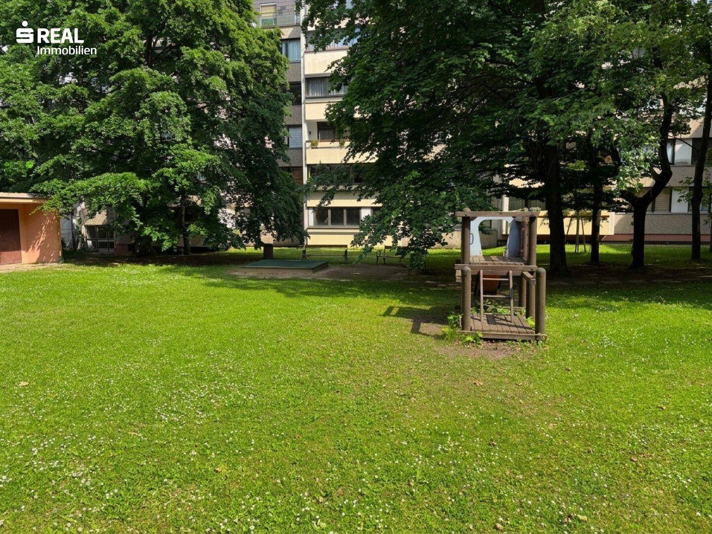 Grüne Rasenfläche mit einem kleinen Spielplatz und Bäumen vor einem Wohngebäude.