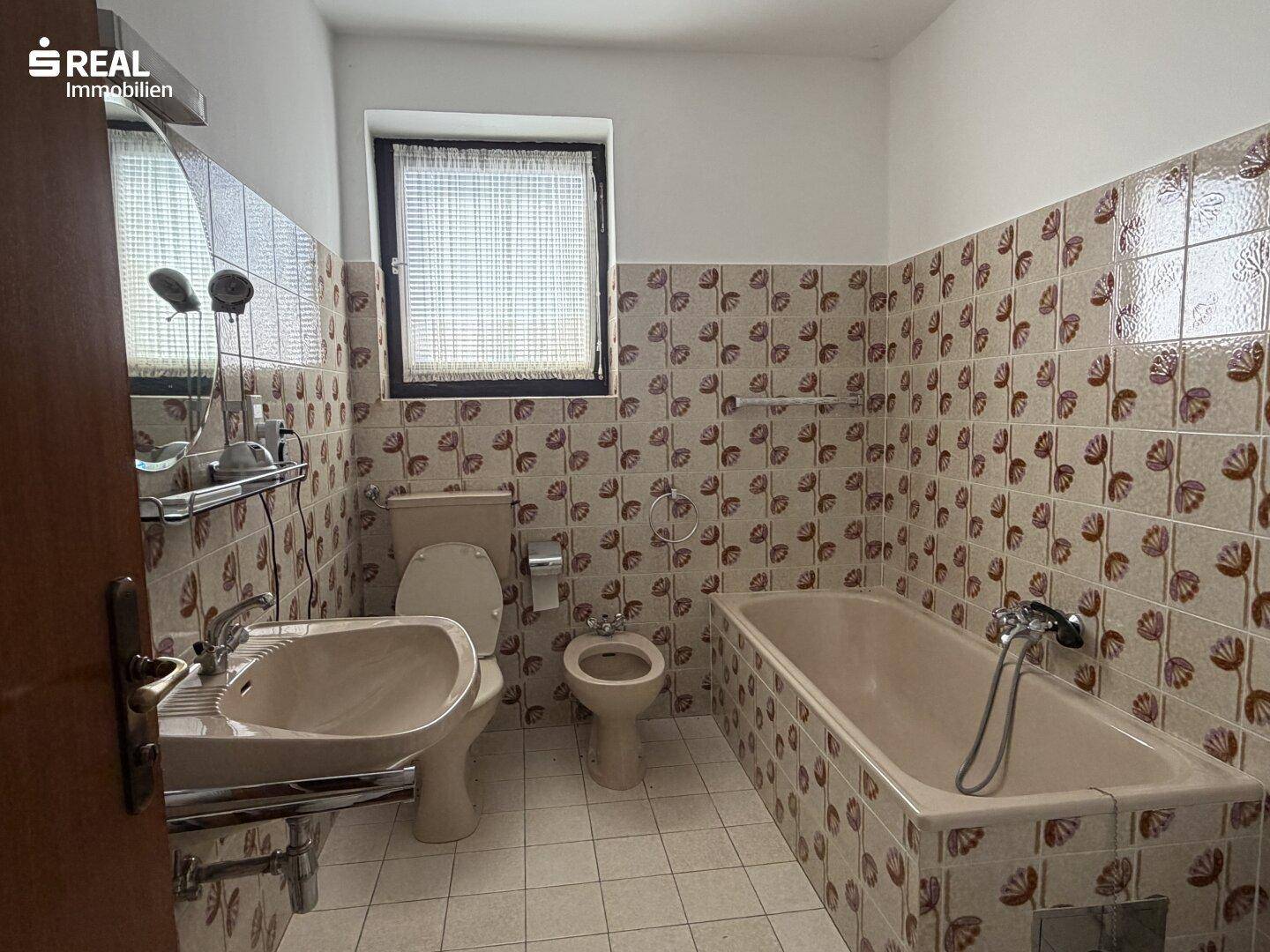Badezimmer mit gemusterten Fliesen, Badewanne, Toilette, Bidet und Fenster für Tageslicht.