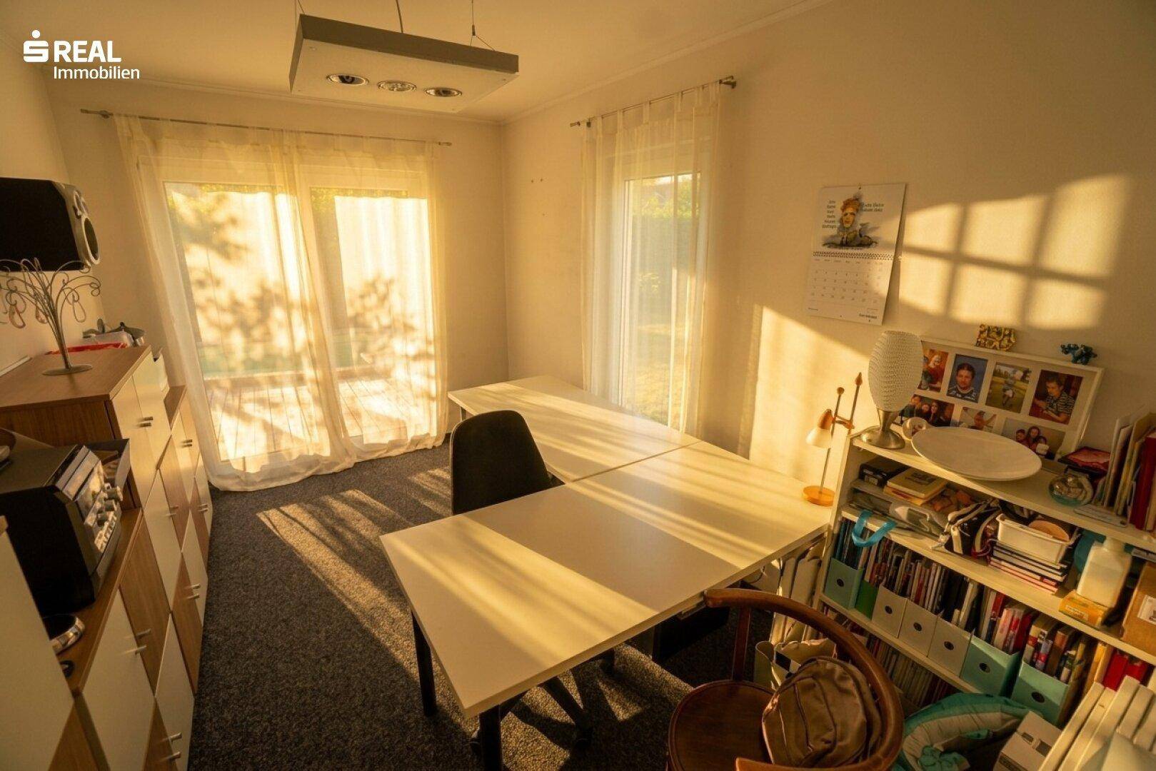 Helles Arbeitszimmer mit zwei Schreibtischen, Teppichboden und großen Fenstern.