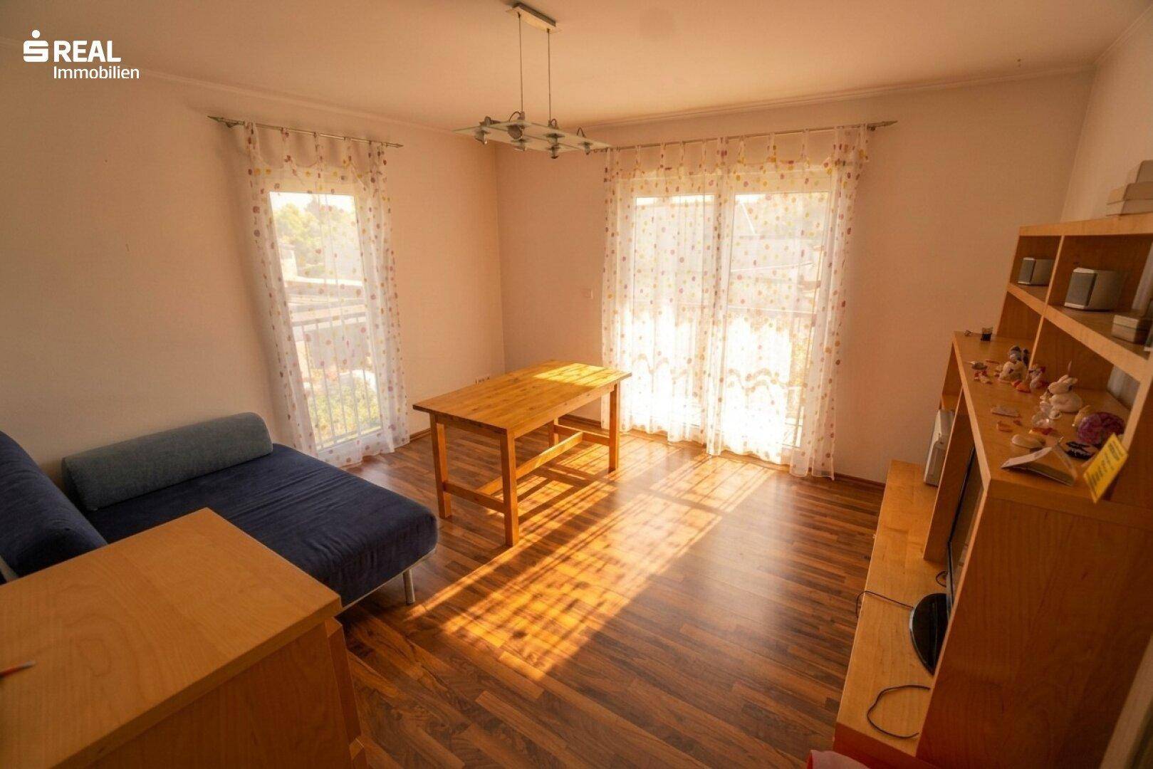 Zimmer mit Holzboden, Schlafsofa und viel Tageslicht durch große Fenster.