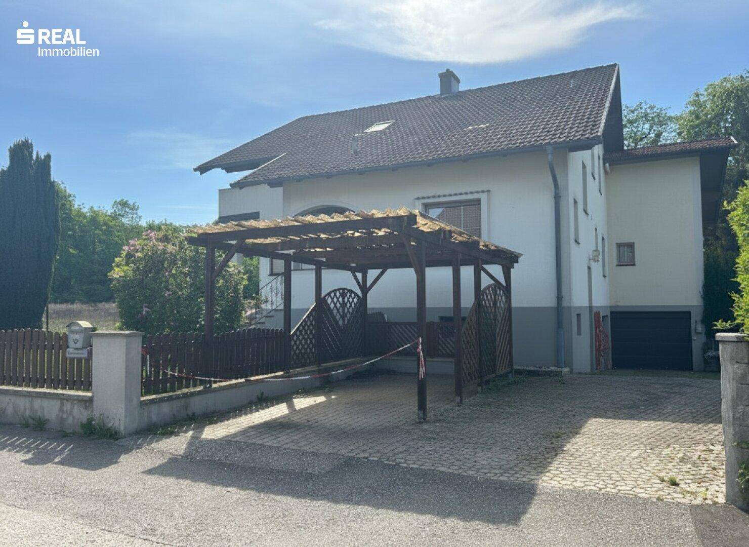 Einfamilienhaus mit überdachtem Carport und Zufahrt, umgeben von Grünflächen.