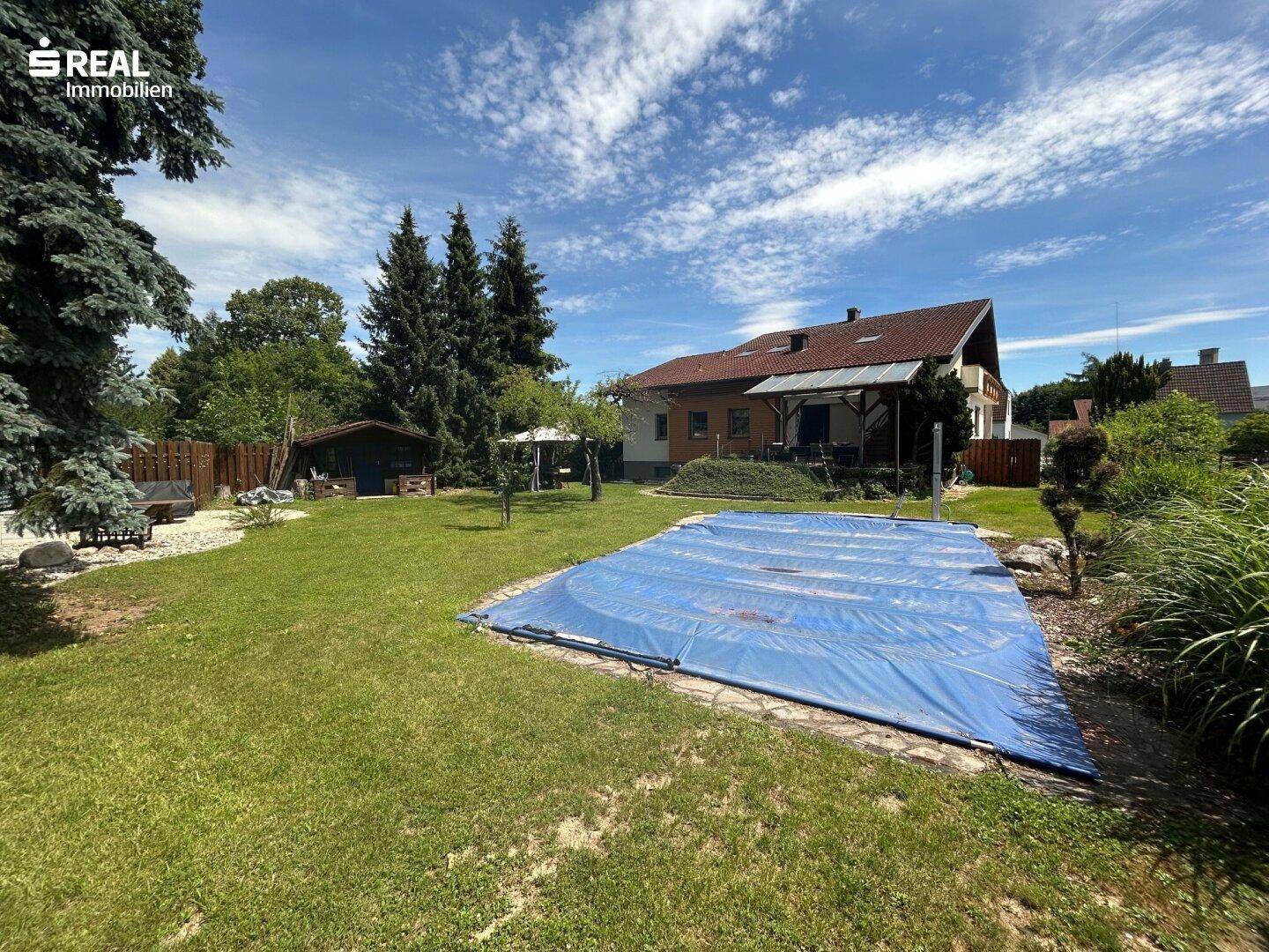 Weitläufiger Garten mit großem, abgedecktem Pool und hohen Bäumen im Hintergrund.