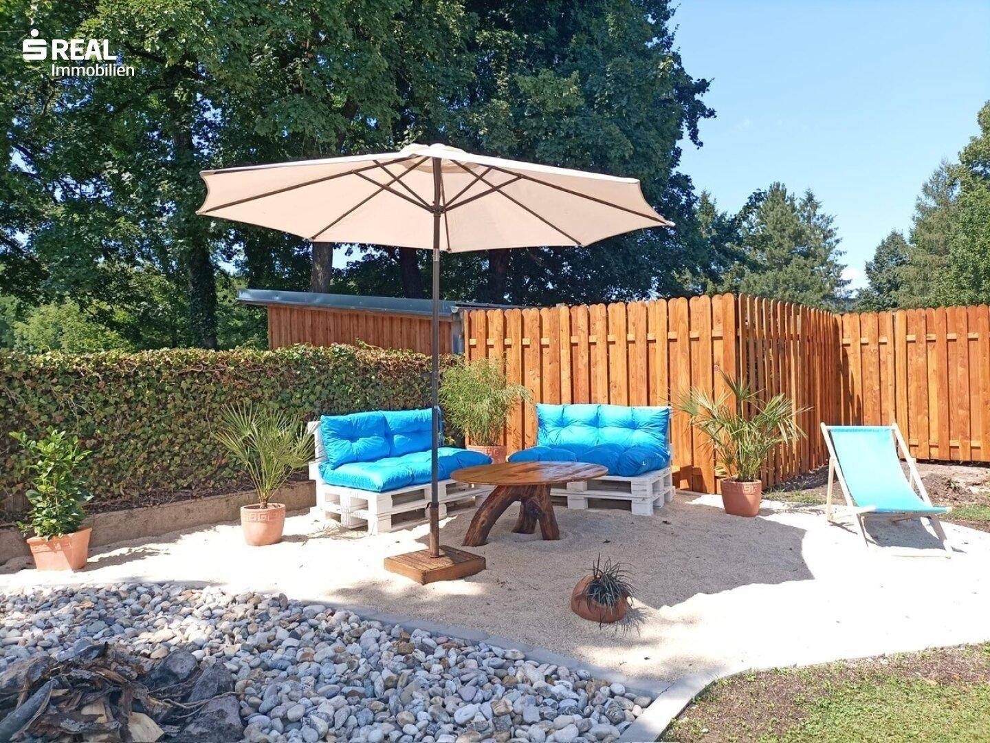Gemütlicher Sitzbereich im Garten mit Sandboden, Palettenmöbeln und Sonnenschirm.