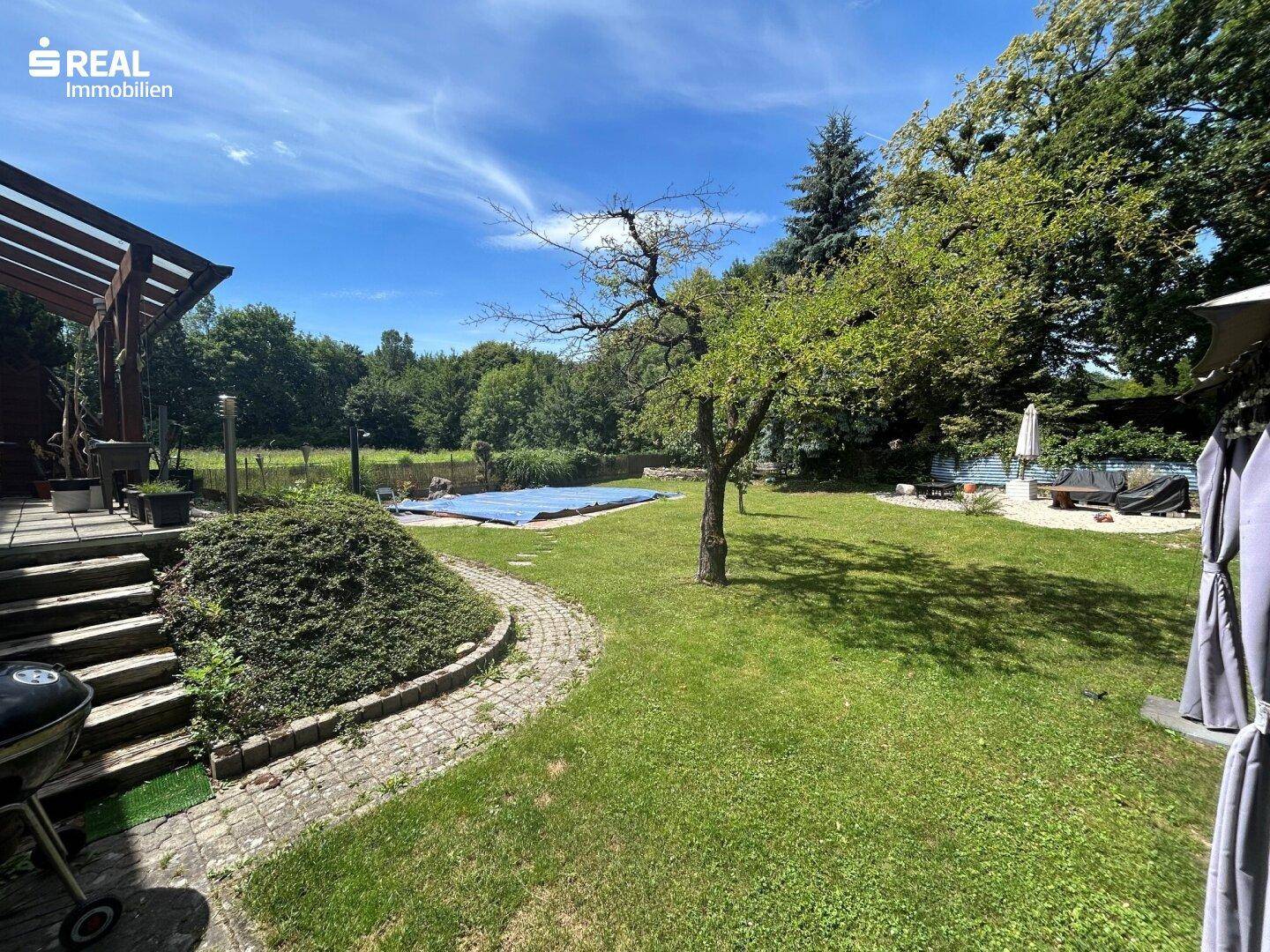 Gepflegter Garten mit Steinweg, altem Baumbestand und Blick auf den Poolbereich.