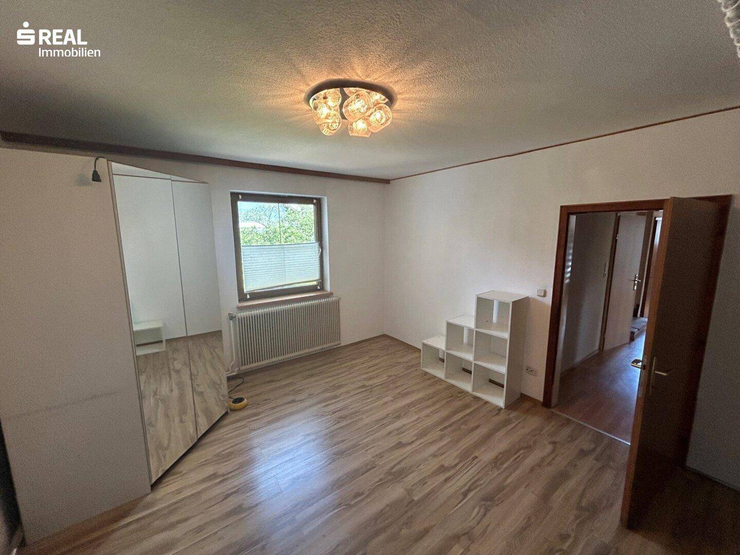 Helles Schlafzimmer mit Laminatboden, Fenster und einfacher Einrichtung.