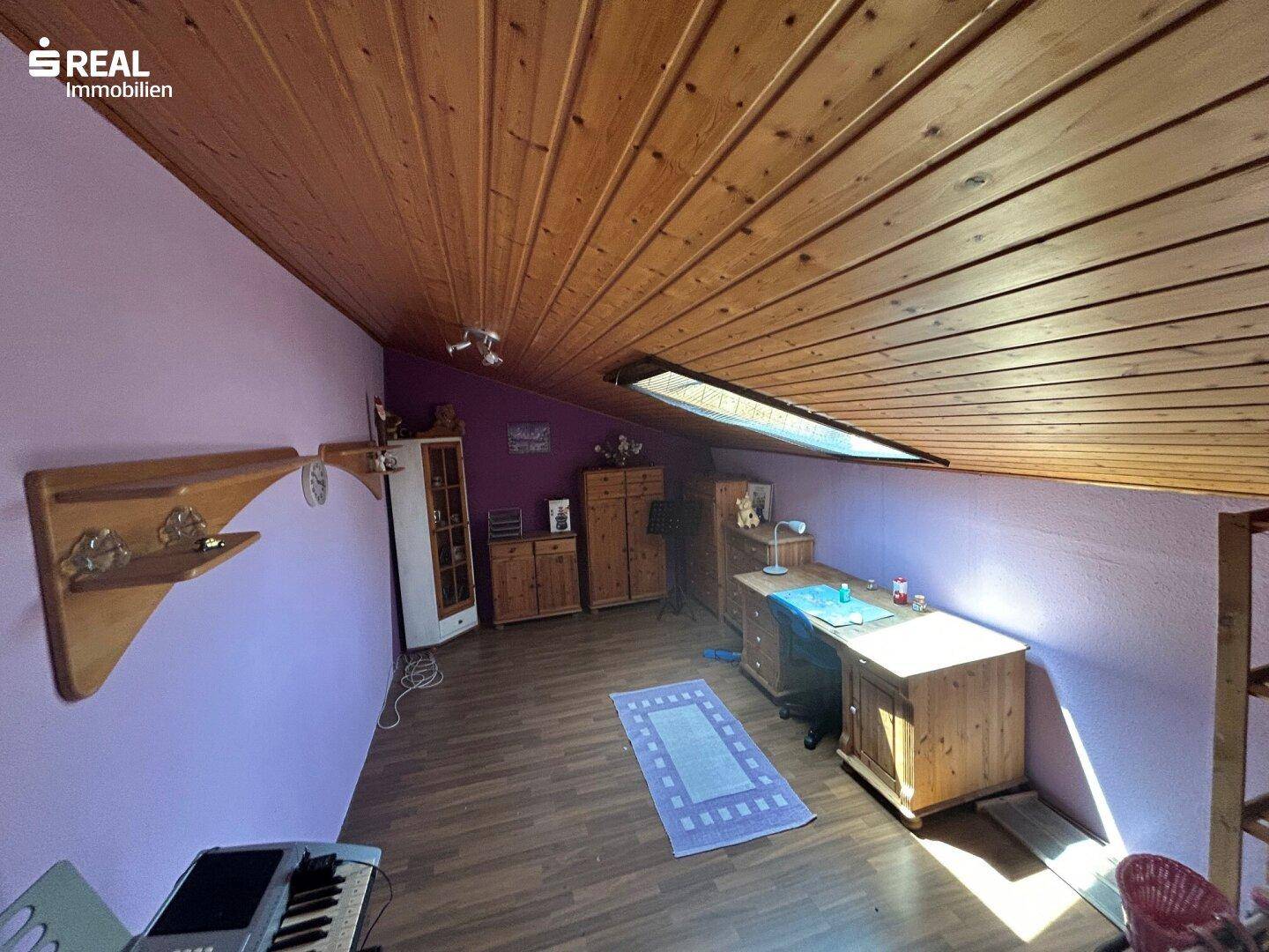 Helles Dachzimmer mit Holzdecke, Schreibtisch, lila Wänden und Dachfenster.