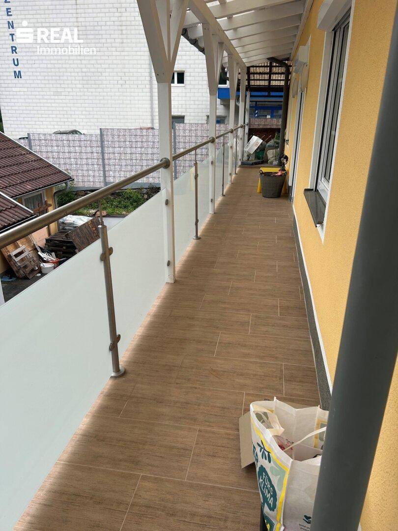Langer, überdachter Balkon mit Holzbodenfliesen und Glasgeländer entlang der Fassade.