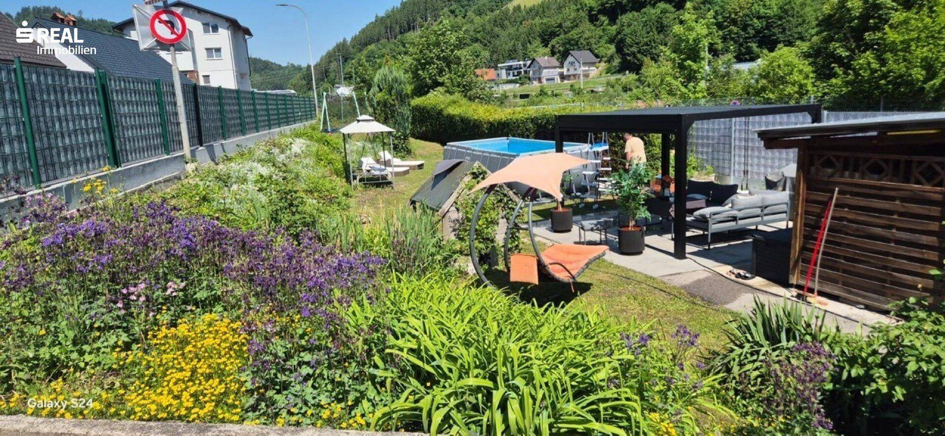 Weitläufiger Garten mit Blumenbeeten, Pool und überdachtem Sitzbereich unter einer Pergola.