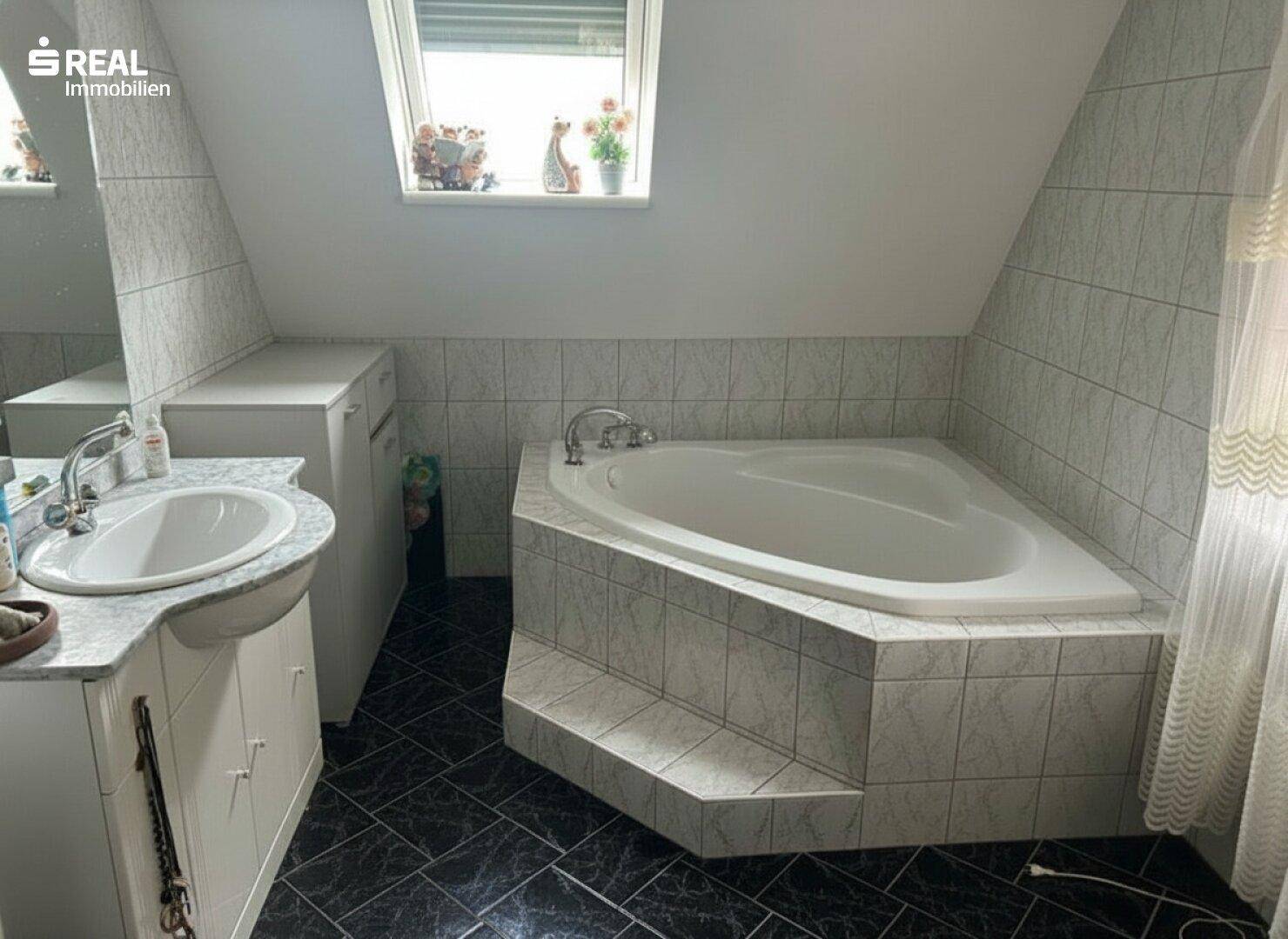Badezimmer im Dachgeschoss mit Eckbadewanne, Waschbecken und dunklen Bodenfliesen.