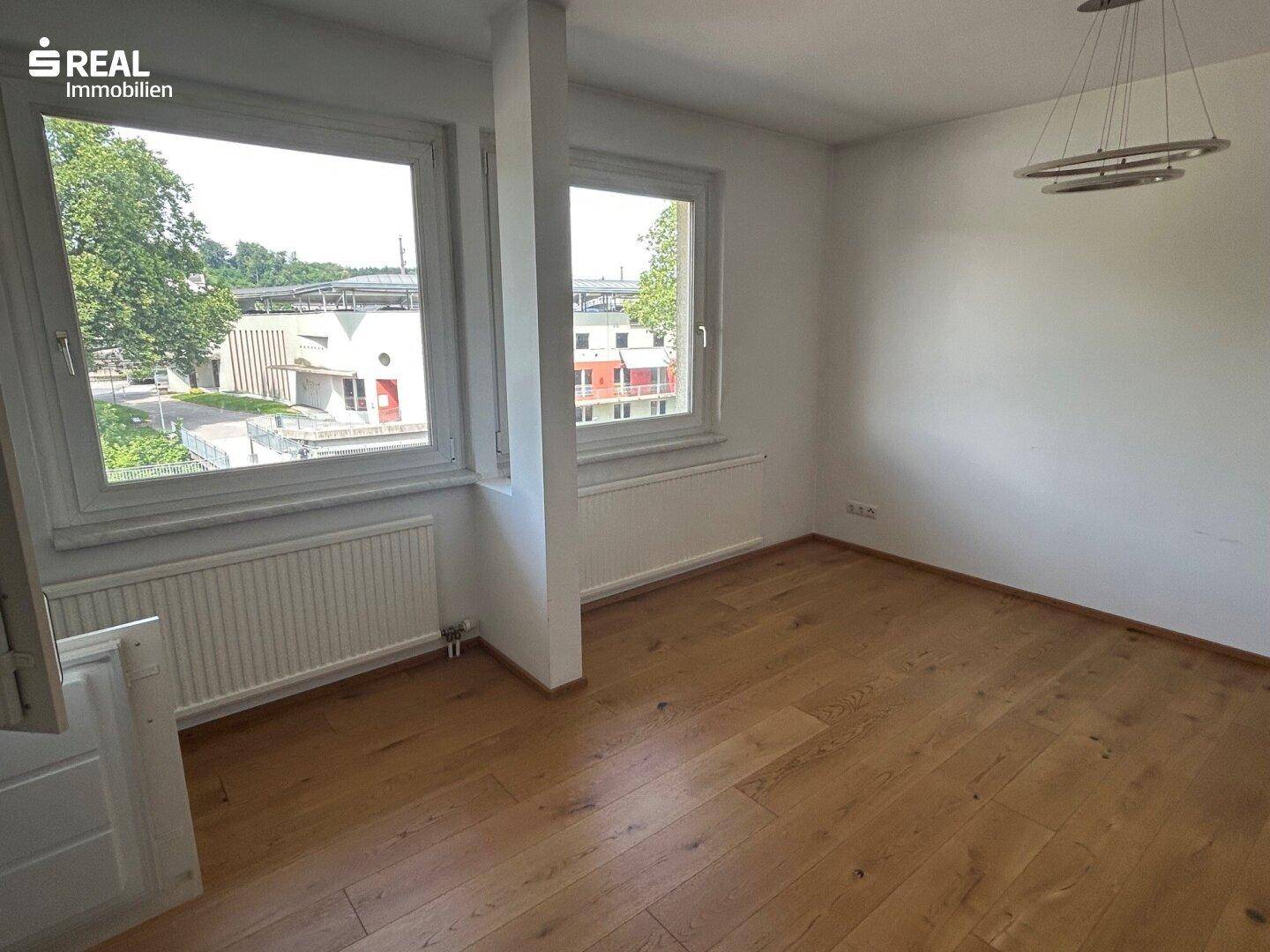 Leerer Wohnraum mit hellem Holzboden, weißen Wänden und zwei großen Fenstern.