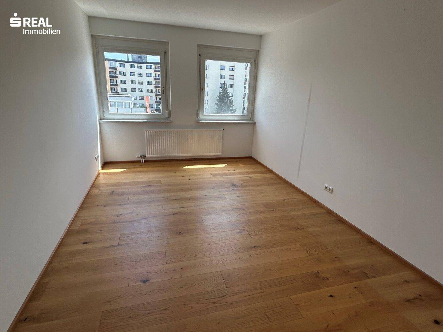 Geräumiges Schlafzimmer mit Holzboden, weißen Wänden und Blick auf umliegende Gebäude und Bäume.