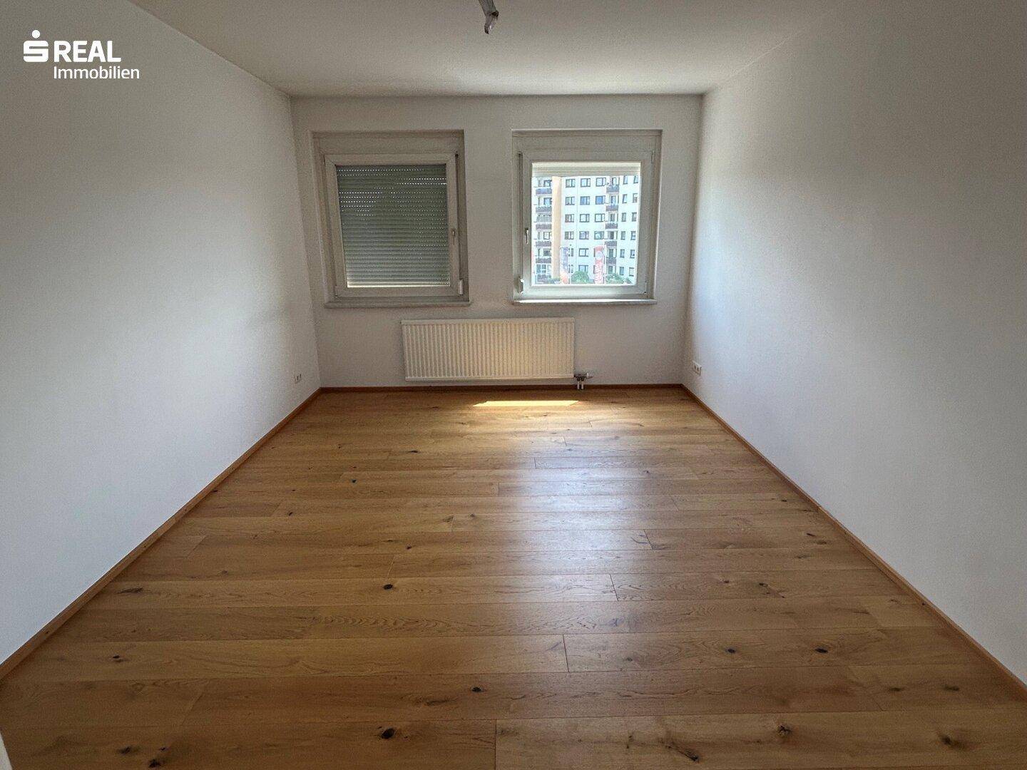 Helles Schlafzimmer mit Holzboden, weißen Wänden und zwei Fenstern, eines davon mit Jalousien.