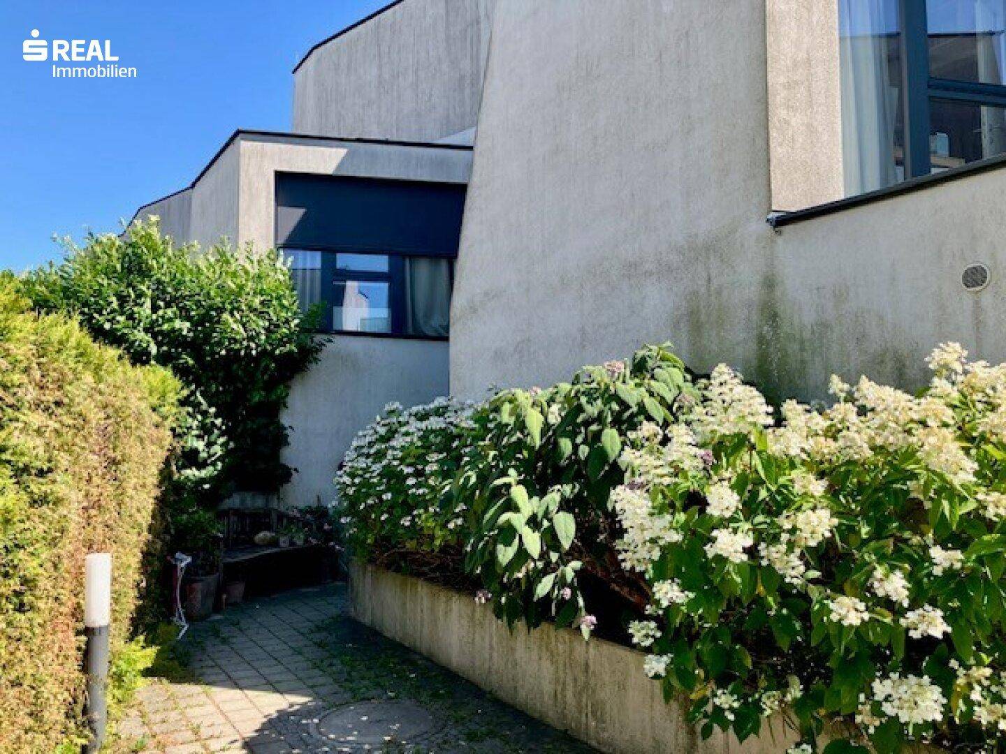 Seitenansicht des Hauses mit gepflastertem Weg und blühenden Sträuchern.