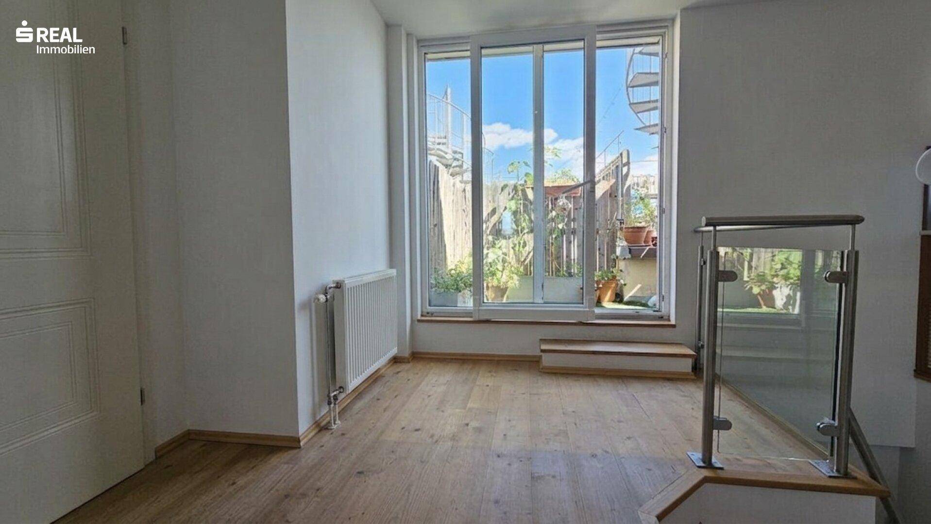 Heller Raum mit Holzboden, großem Fenster und Zugang zu einer Außenterrasse über eine Treppe.