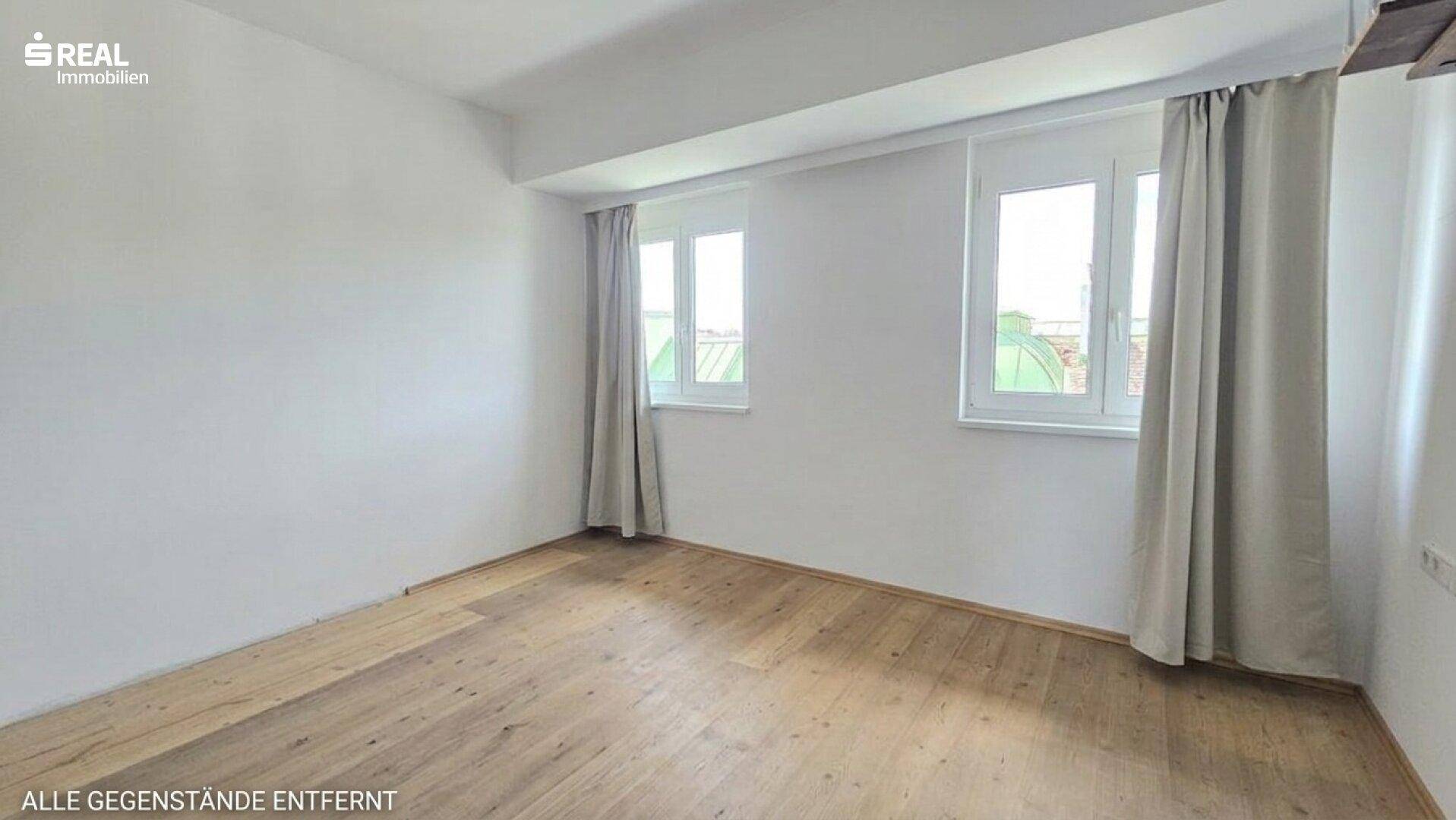 Helles, leeres Zimmer mit Holzboden, weißen Wänden und zwei Fenstern mit Vorhängen.