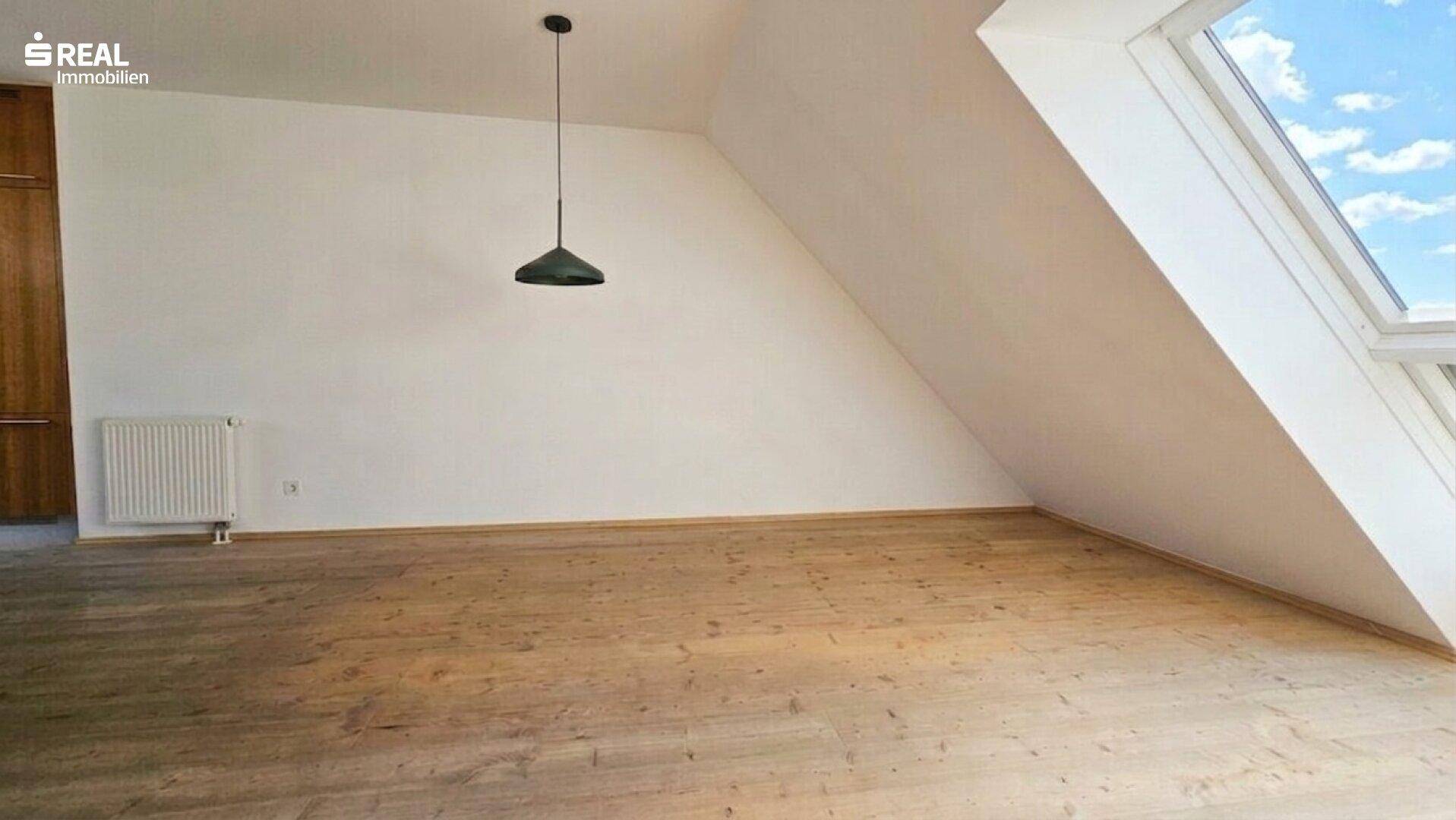 Geräumiges Dachzimmer mit Holzboden, weißen Wänden, Dachfenster und schrägen Decken.