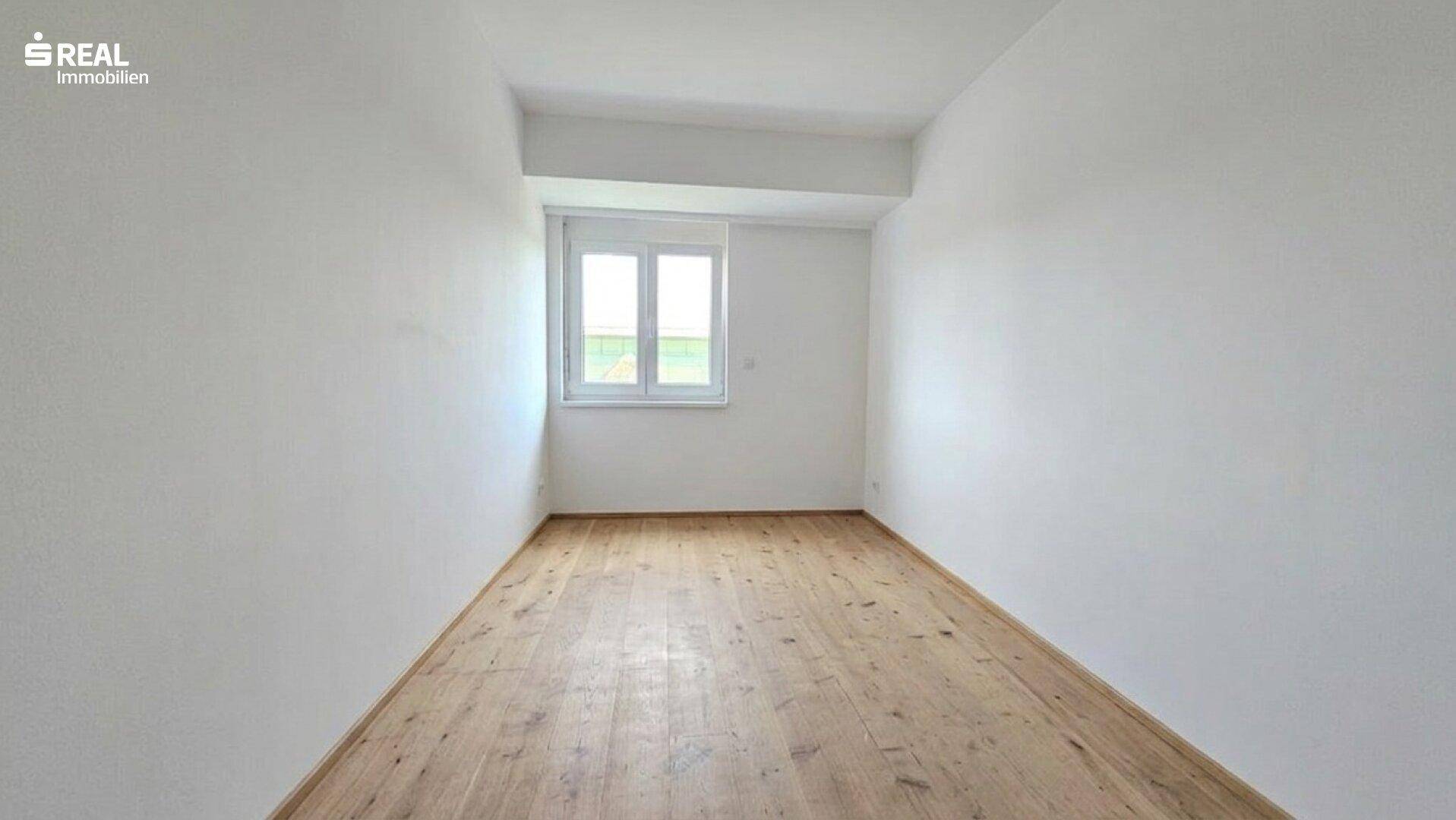 Schlichtes, leeres Zimmer mit Holzboden, weißen Wänden und einem Fenster für natürliches Licht.