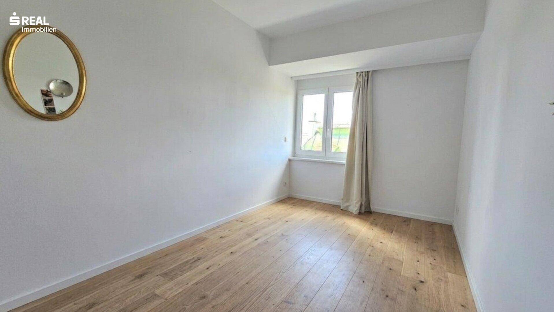 Helles, leeres Zimmer mit Holzboden, weiß gestrichenen Wänden und einem Fenster.