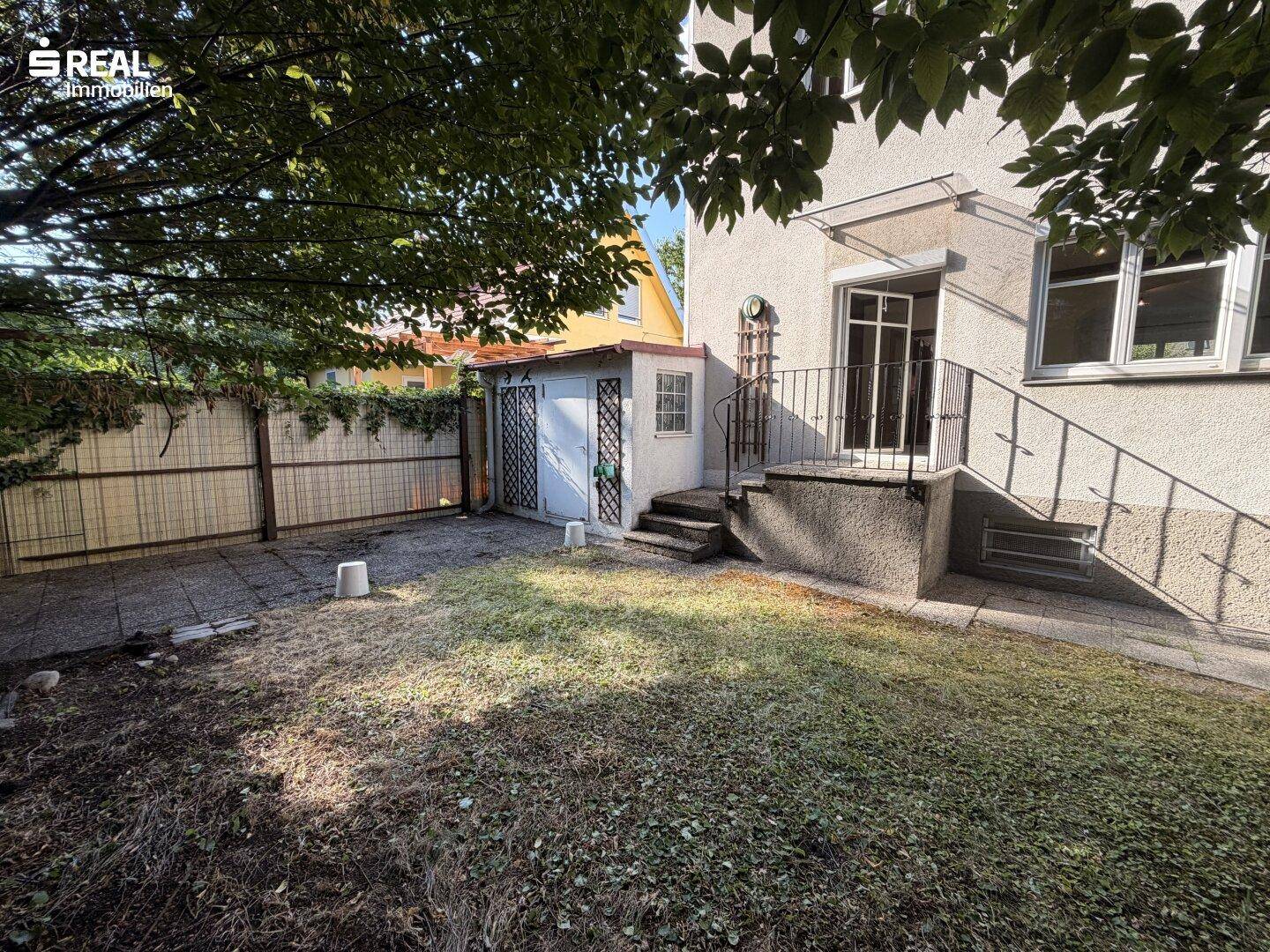 Sonnige Gartenfläche hinter dem Haus mit Nebengebäude und Treppe zum Eingang.