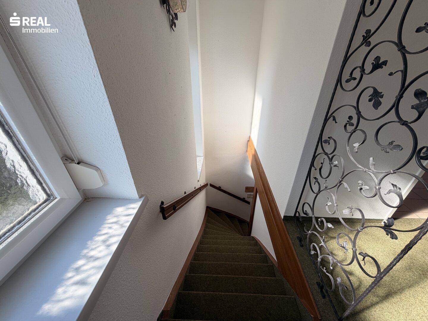 Treppenhaus mit Teppichstufen, schmiedeeisernem Geländer und natürlichem Lichteinfall.