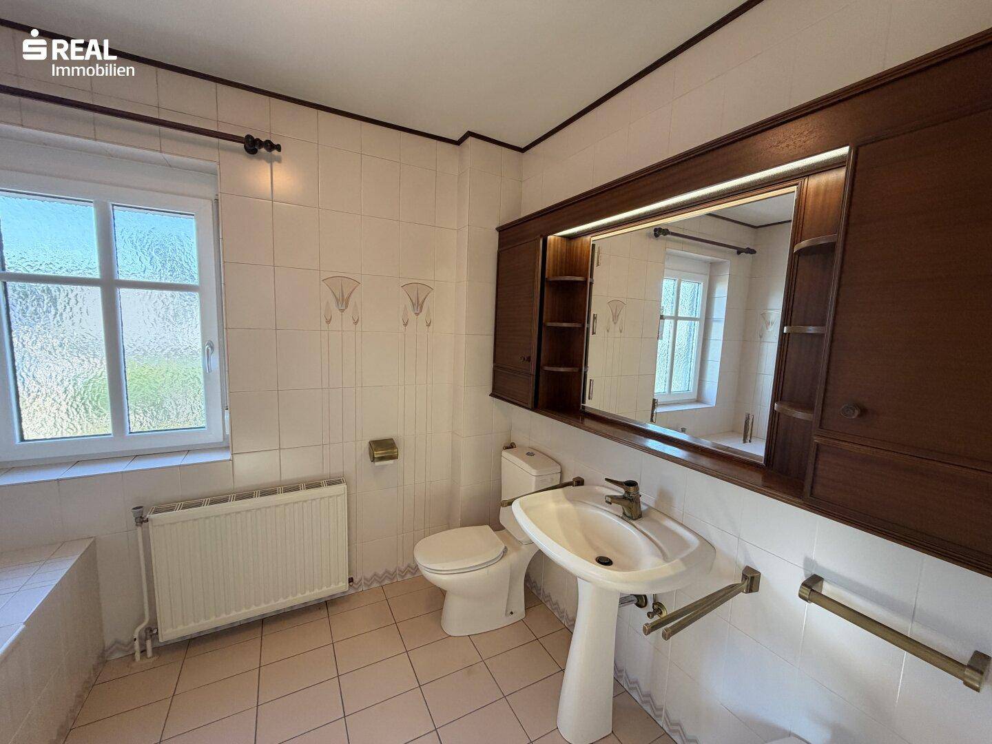 Badezimmer mit Fliesenboden, Fenster, Waschtisch und Spiegelschrank.